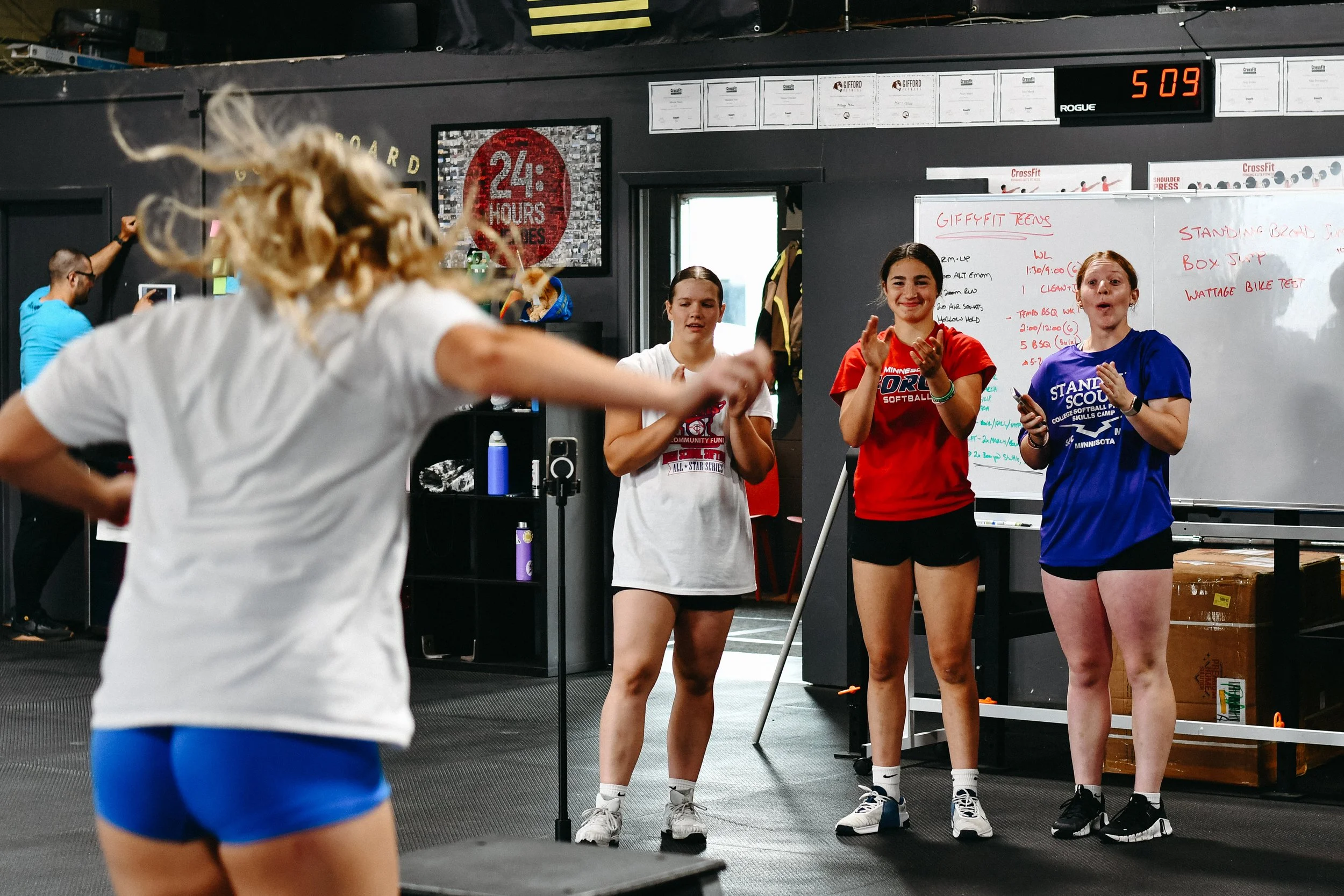 Four young women in athletic clothing inside a gym, with one woman in the foreground jumping onto a tall box.