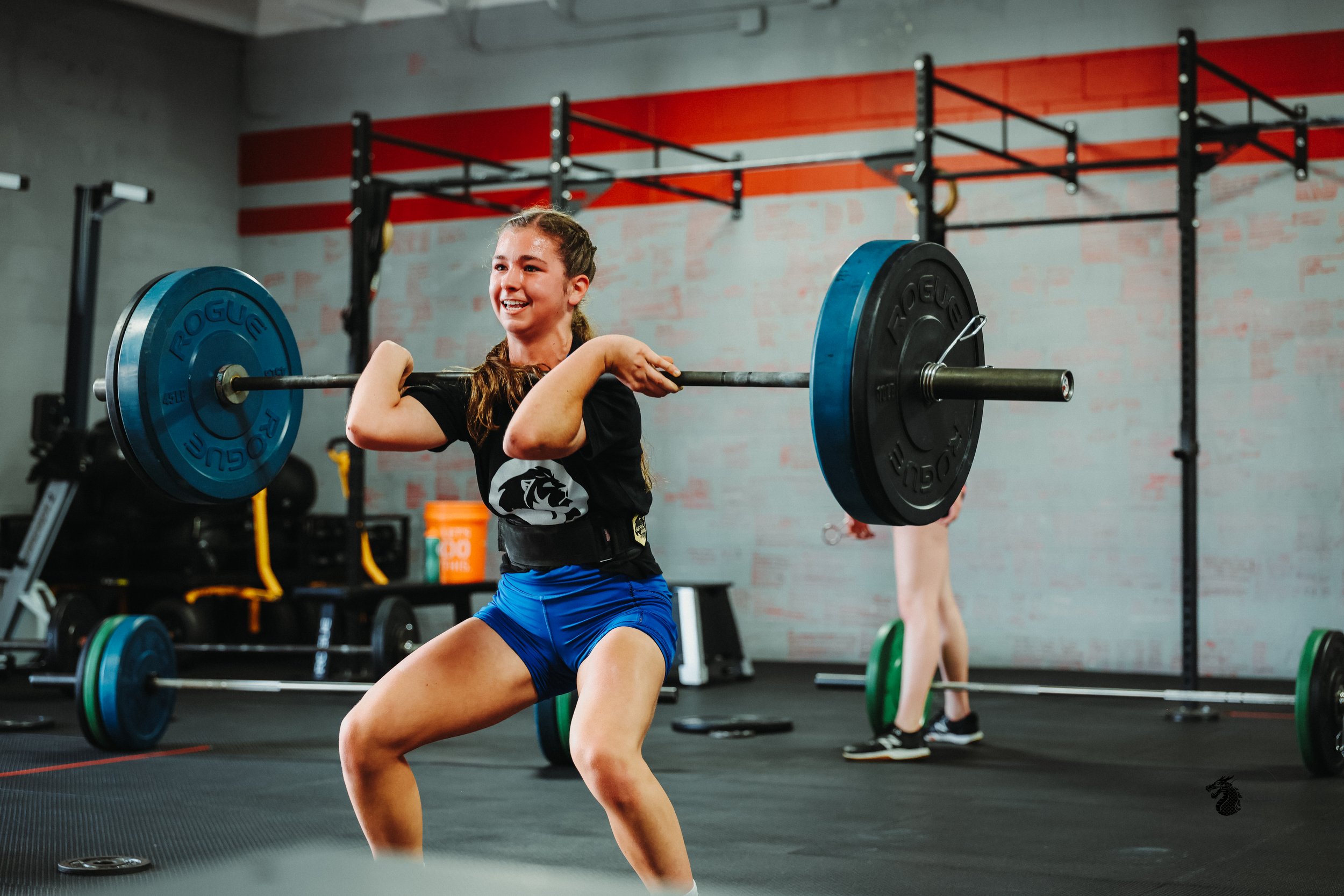 A young woman lifts a barbell with blue and black weights during a workout at a gym.
