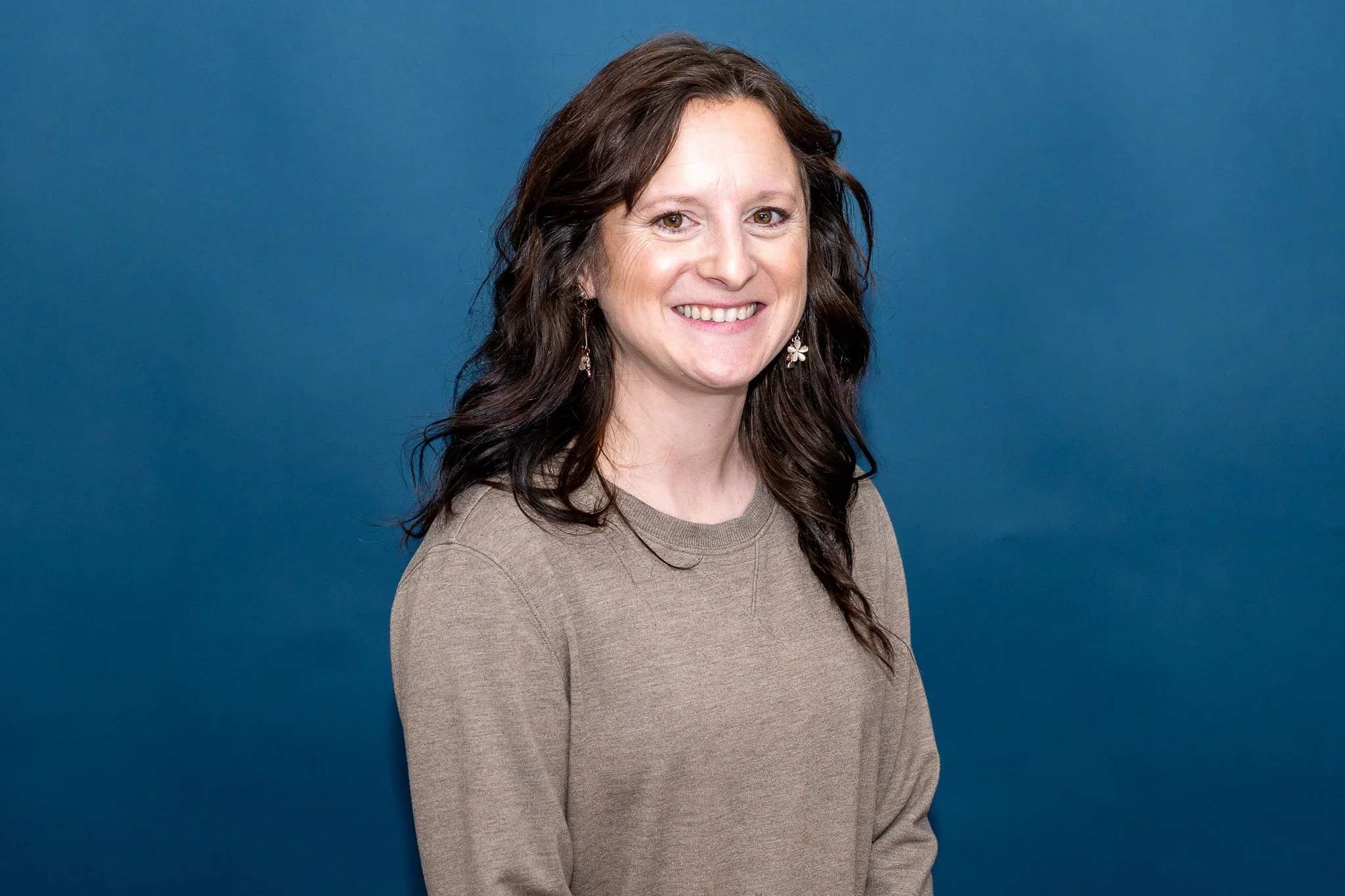 A woman with shoulder-length wavy light brown hair, wearing glasses and a pink blazer, smiling against a blue background.