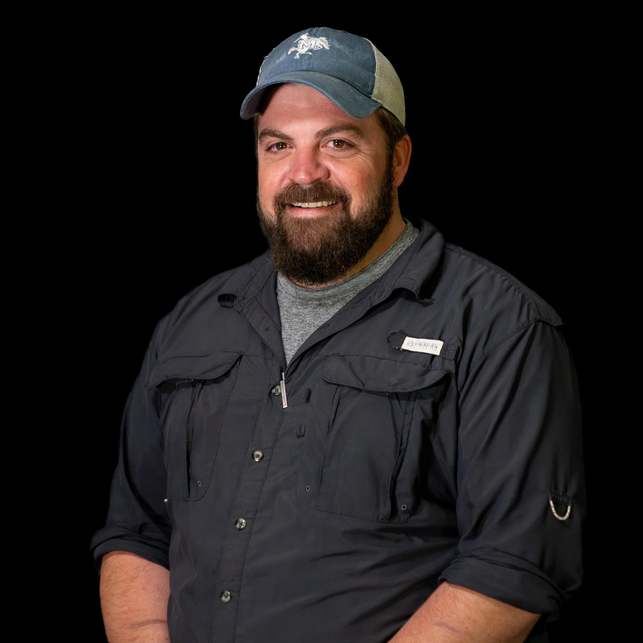 Young man with long brown hair, wearing a black beanie and a gray uniform shirt with a name tag, smiling against a blue background.