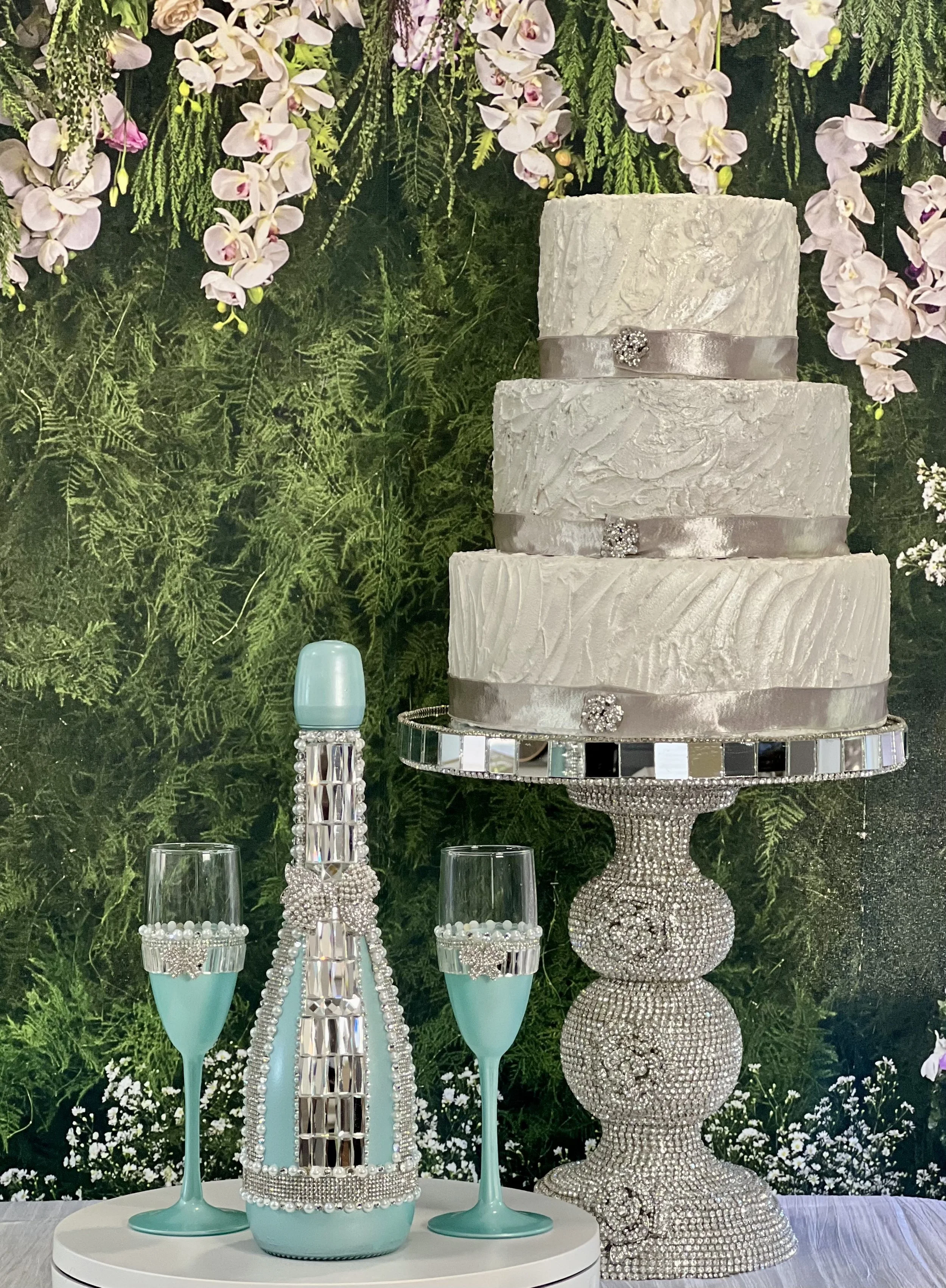 A multi-tiered white wedding cake with textured icing and decorative brooches, set on a sparkling, jeweled cake stand. Beside it are two champagne flutes with decorative bands and a light blue bottle with jeweled embellishments. The background features pink orchids and greenery.