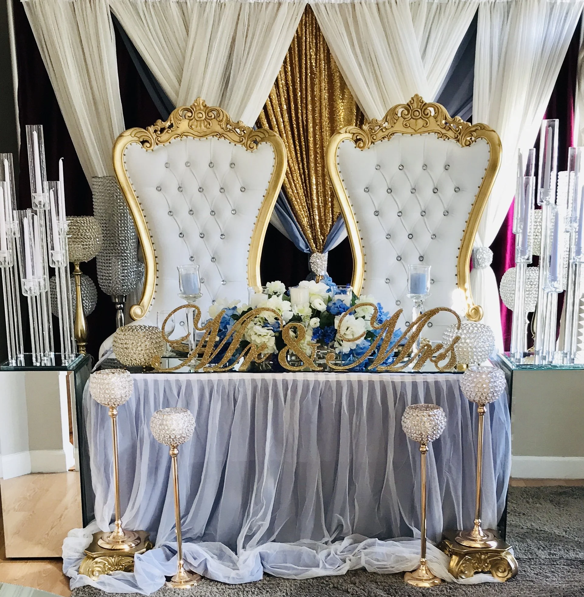 Elegant wedding table setup with our white/gold throne chairs. Bride & Groom table decorated with custom rhinestone "Mr & Mrs" sign, set against a backdrop of draped white and gold curtains.