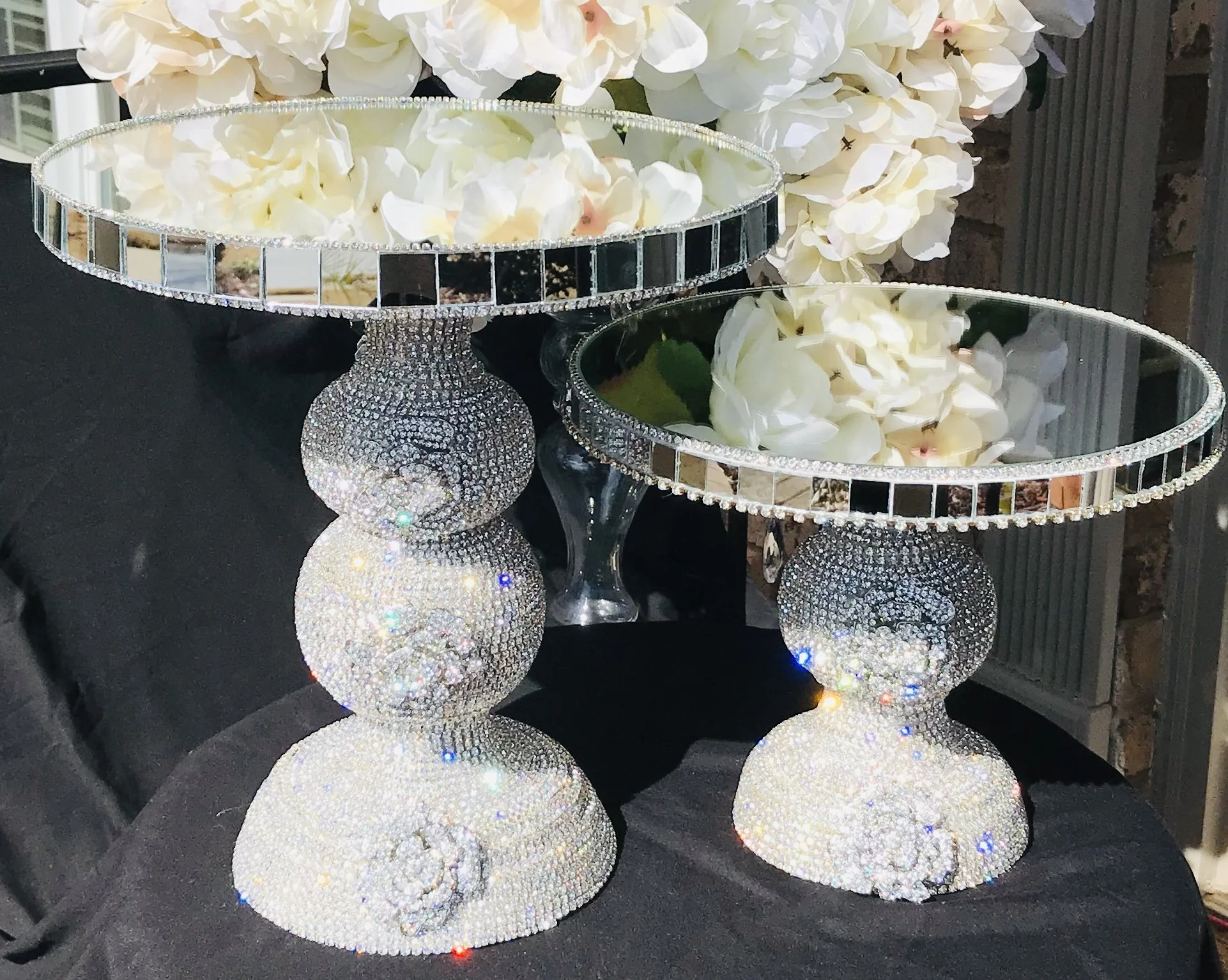 Two decorative tables with reflective mirrored rims and bases encrusted with rhinestones, topped with white hydrangea flowers, placed on a black cloth surface.