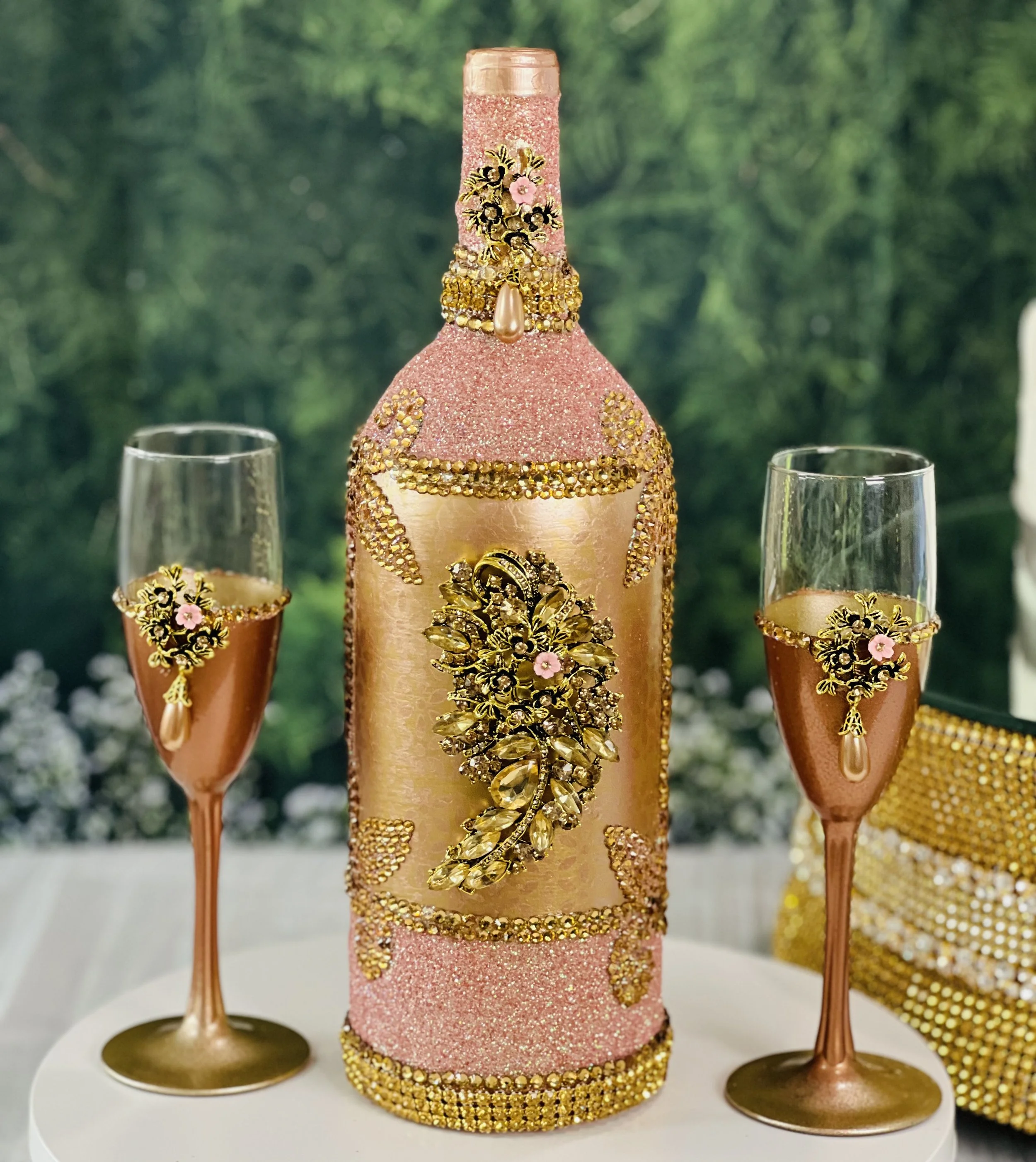 Decorative pink champagne bottle with gold embellishments and matching glasses on a white table.