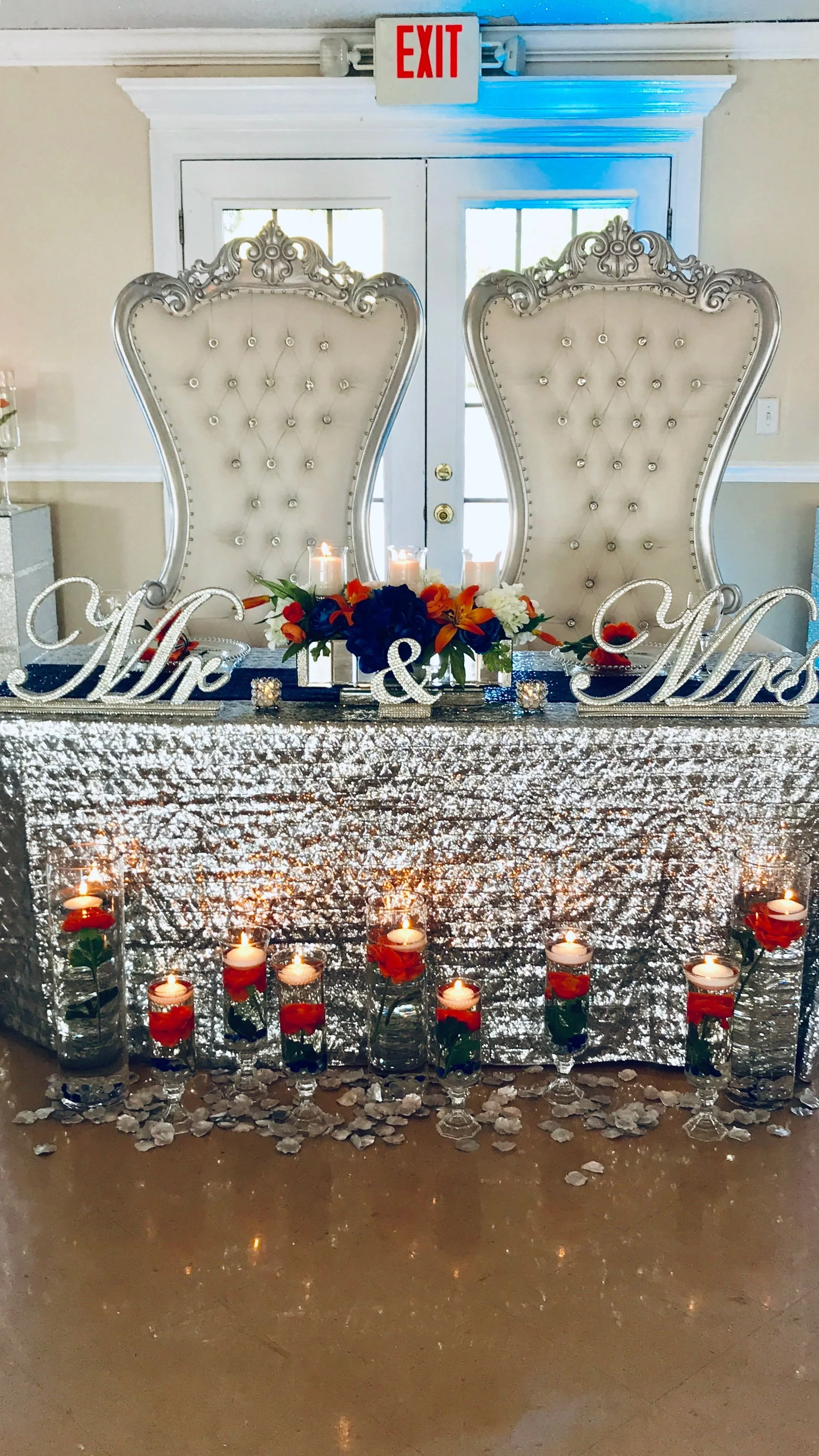 Wedding sweetheart table with two high-backed ornate chairs, silver sequin tablecloth, floral centerpiece, Mr. & Mrs. signs, candles, and floating candles with roses inside glass vases, in front of white double doors with an exit sign above.