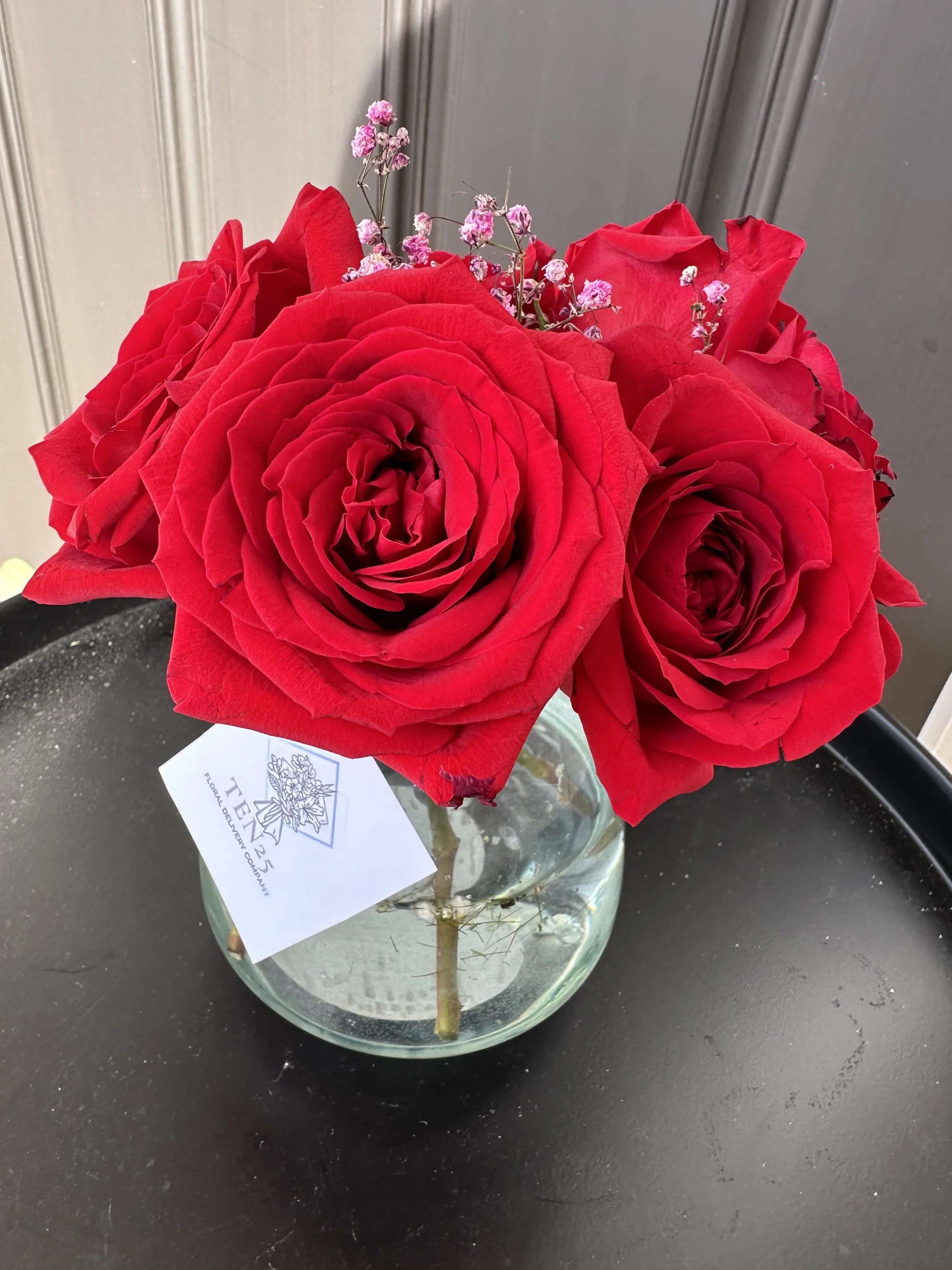 A bouquet of red roses and pink baby's breath in a round glass vase on a black surface.