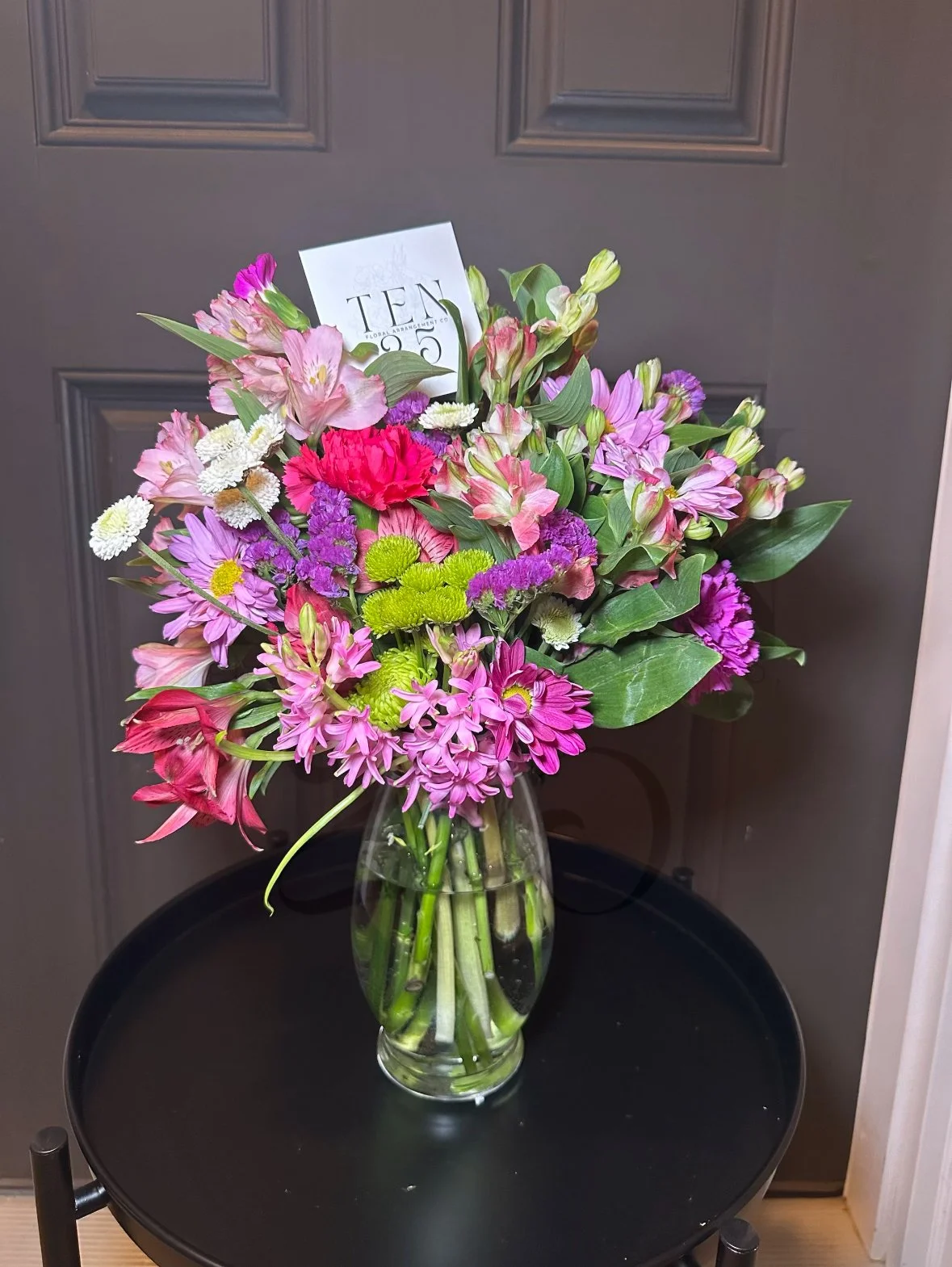 A bouquet of pink, purple, white, and green flowers in a glass vase on a round black table, set against a gray paneled door.