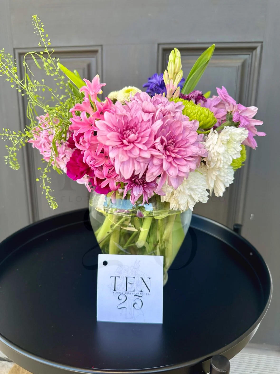 A bouquet of pink, white, purple, and green flowers in a glass vase on a black round tray with a white card labeled 'TEN 25' in front of a gray wall.