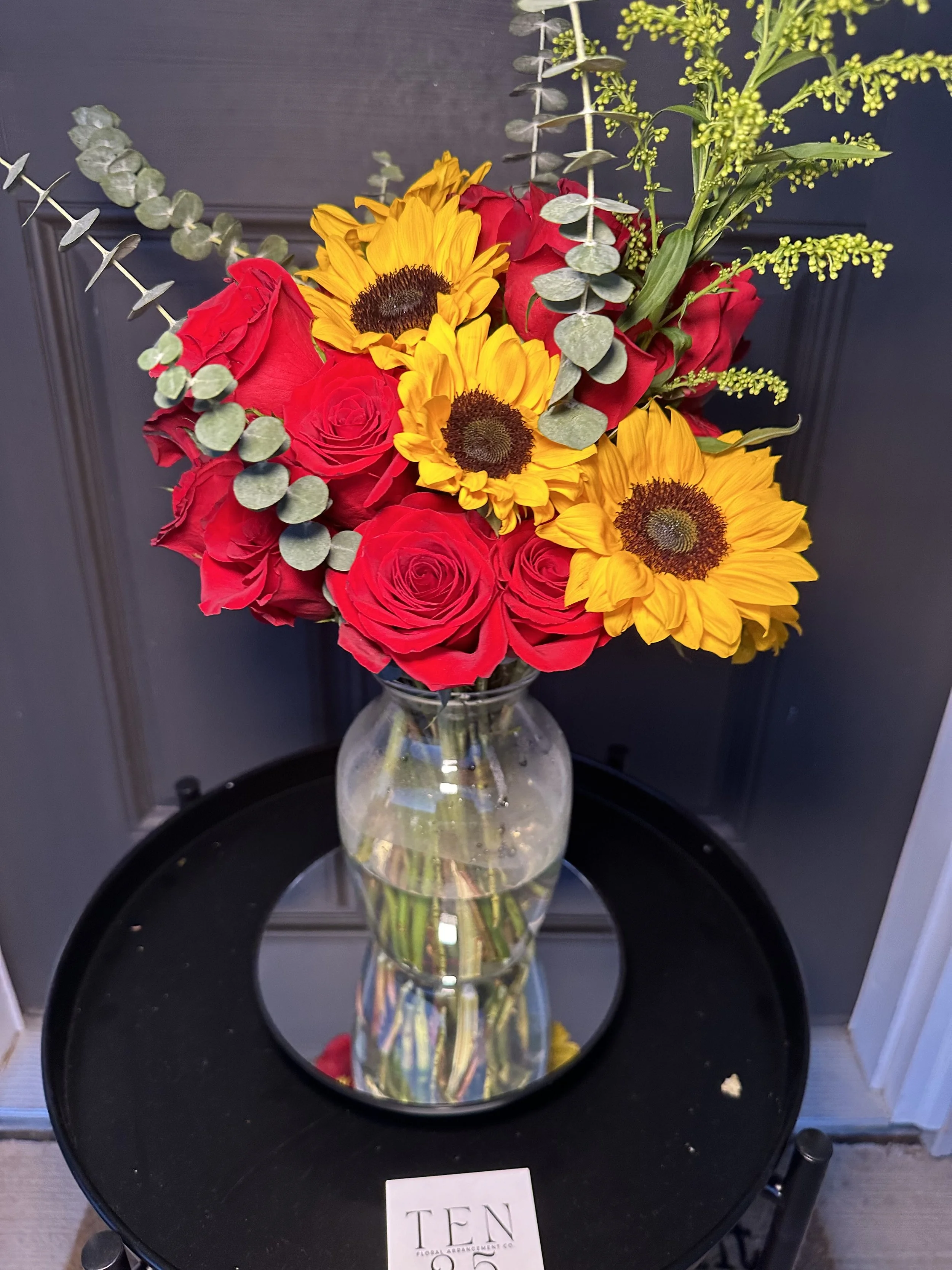 A bouquet of red roses, yellow sunflowers, and green foliage in a glass vase on a black round table.