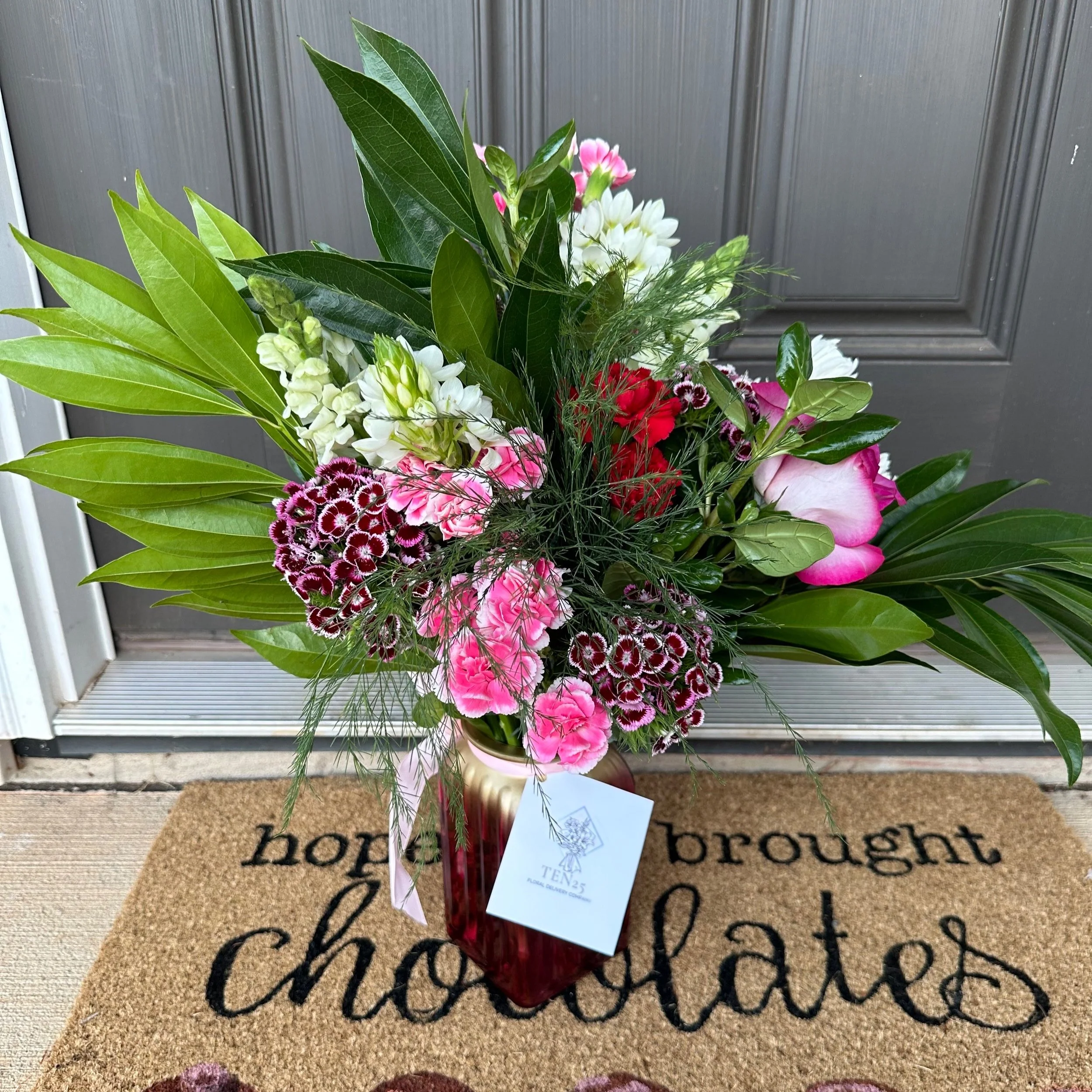 A colorful flower arrangement in a red vase placed on a doormat that says "hope brought chocolates" in front of a gray door.