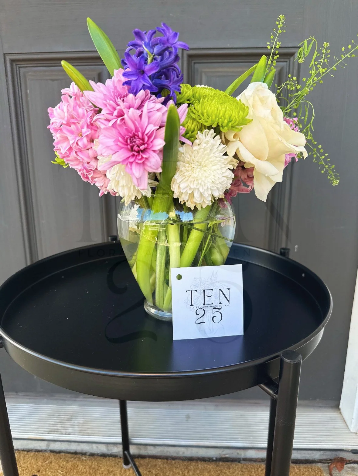 A glass vase with a colorful flower arrangement on a black round side table. The bouquet includes pink, purple, white, and green flowers with green leaves. There is a small card with the text 'TEN 25' leaning against the vase. The background shows a 