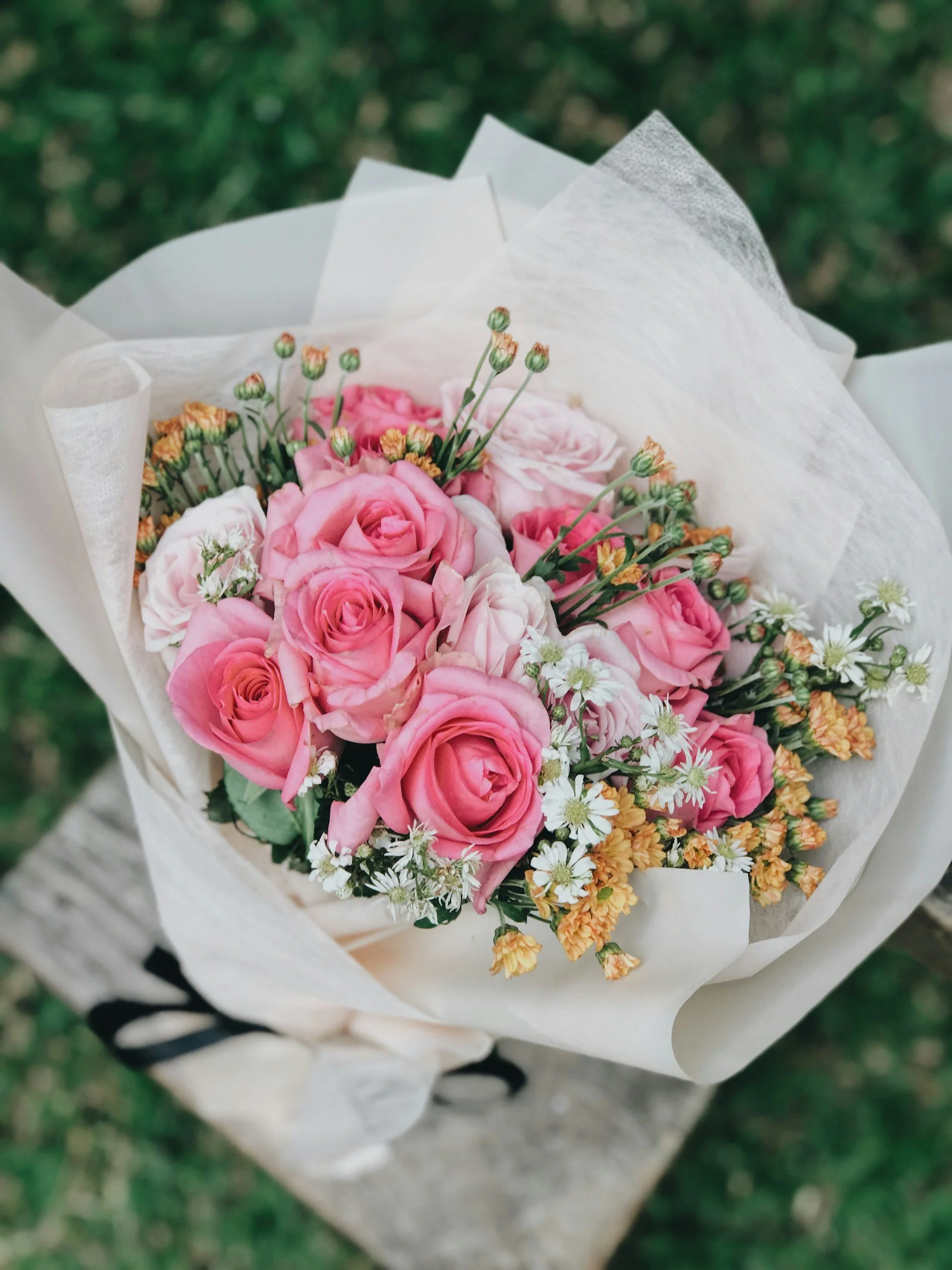 A bouquet of pink roses, white daisies, and orange filler flowers wrapped in white paper.