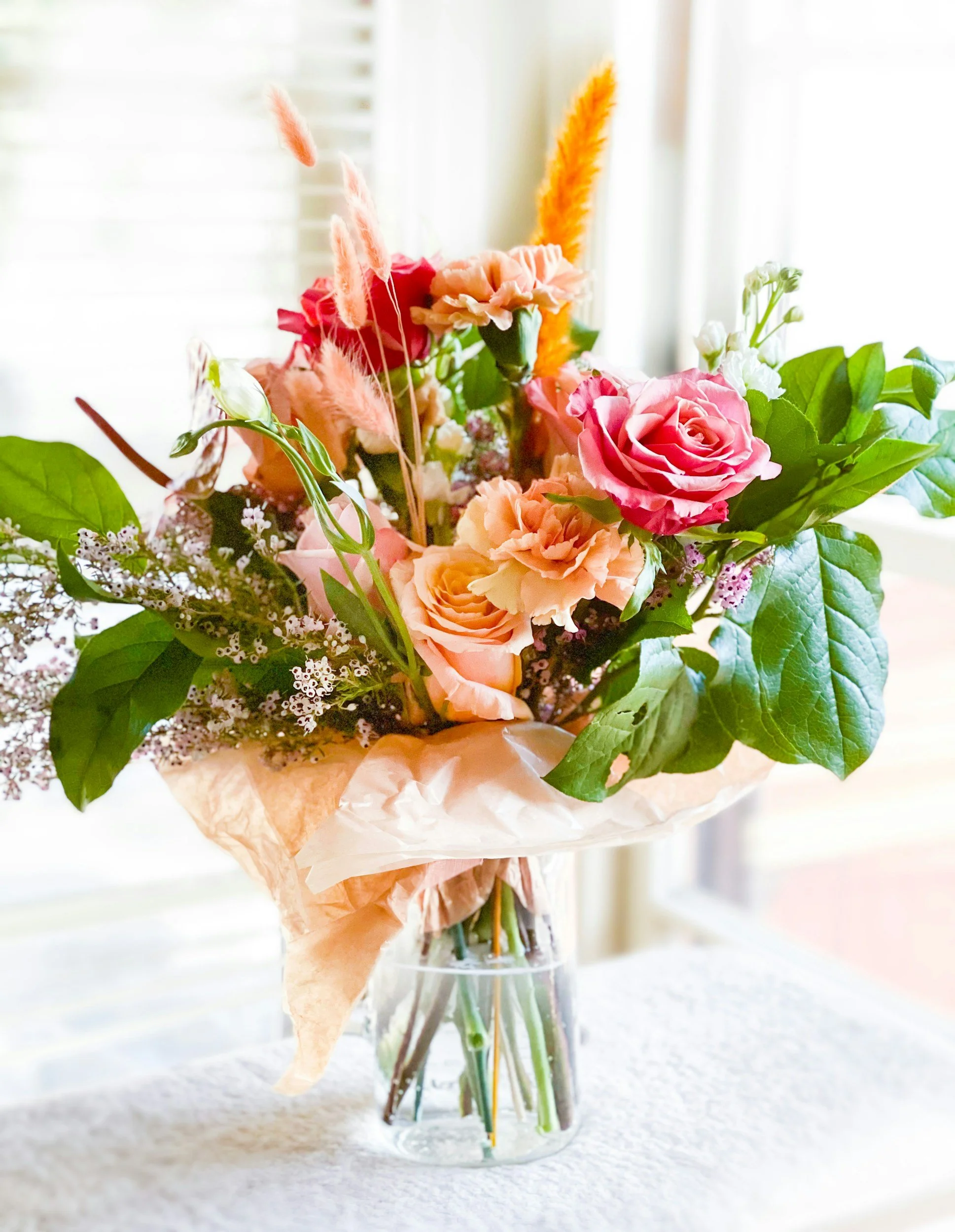 A bouquet of pink and peach roses, green leaves, and pink and orange fluffy dried flowers in a clear glass vase.