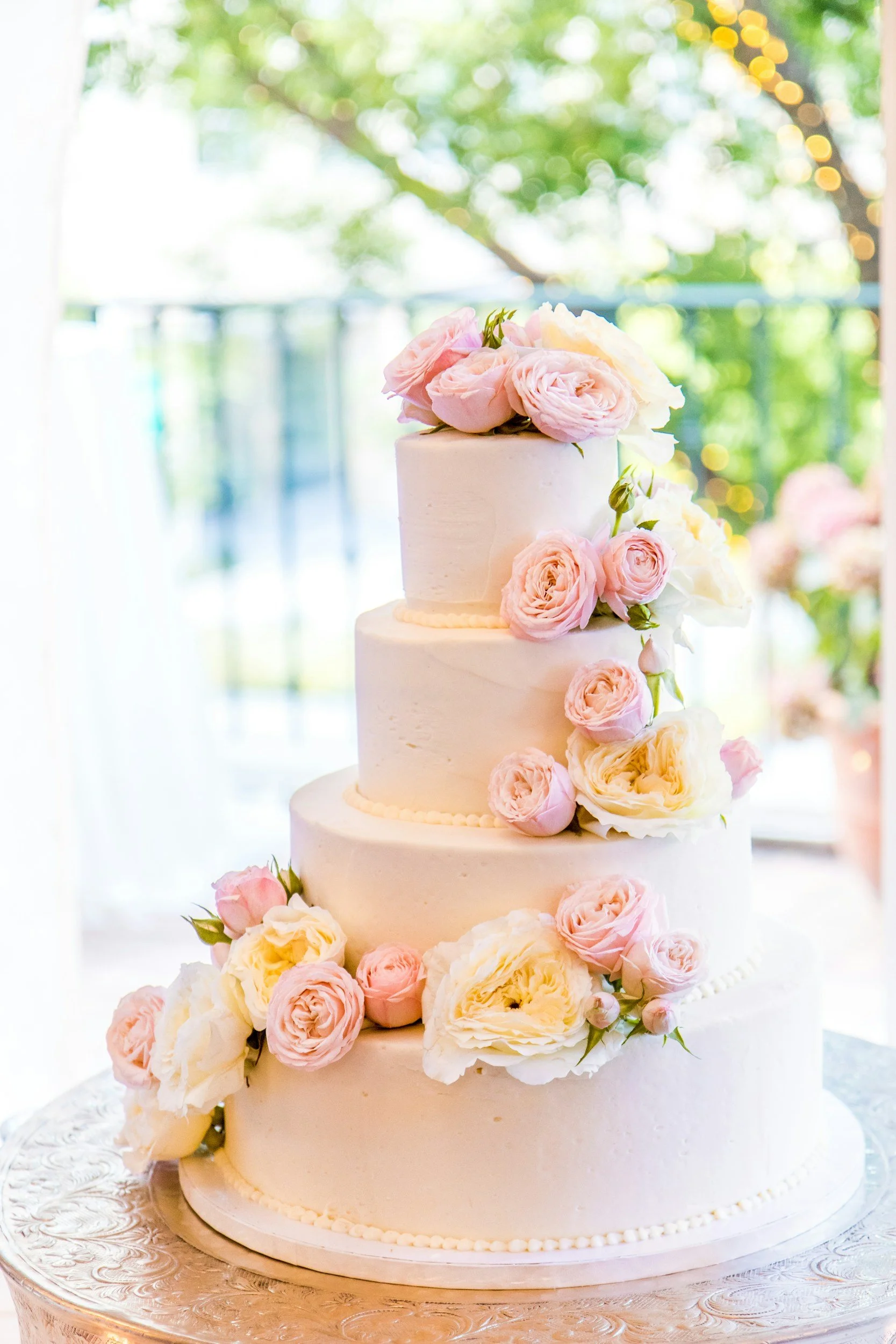 A three-tiered white wedding cake decorated with pink and white roses, placed on a silver cake stand with intricate patterns, set against a backdrop of greenery and bokeh lights.