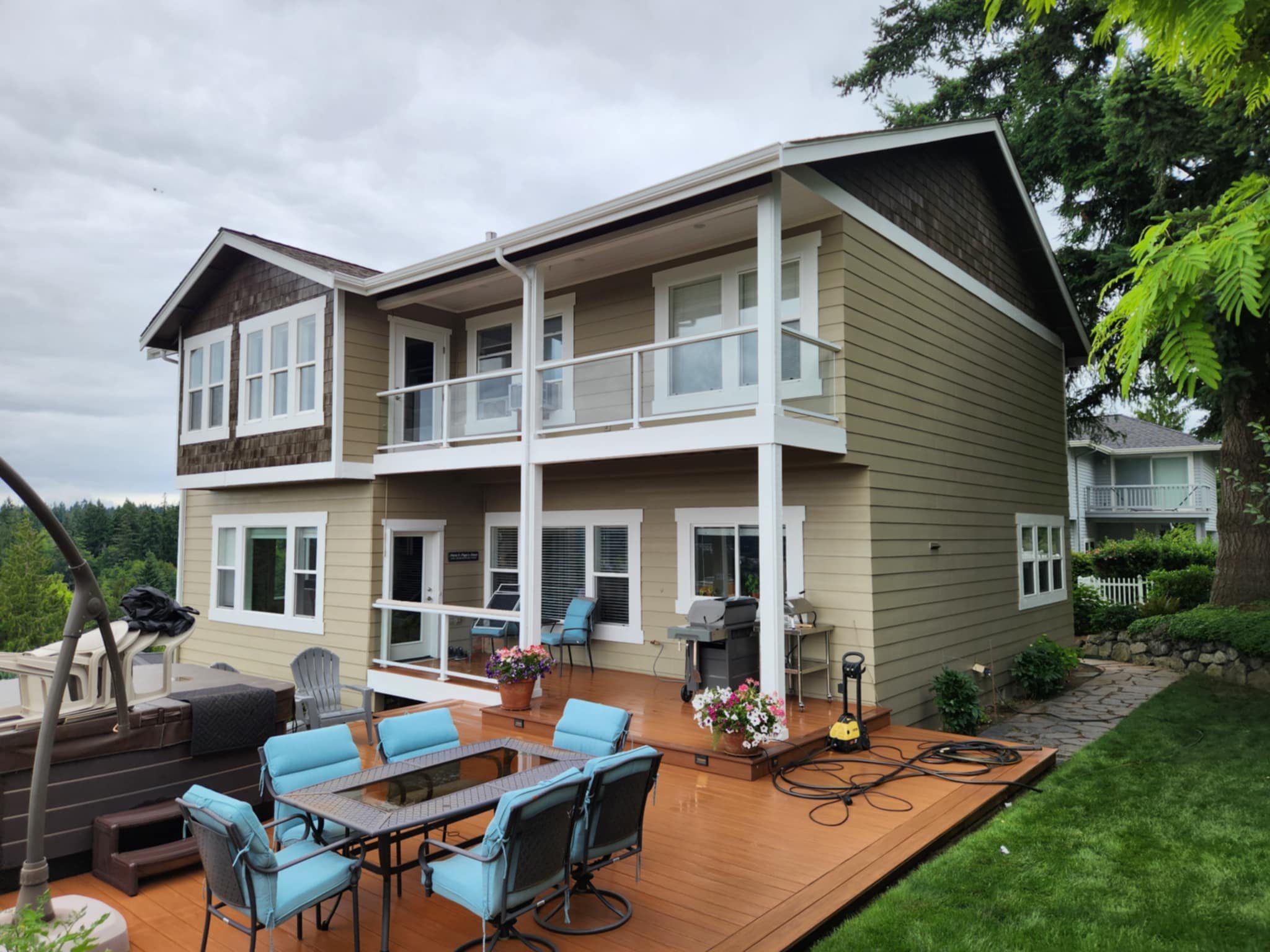 The image shows the backyard of a house with a large wooden deck, outdoor furniture including a table with blue cushions, potted flowers, and a grill. The house has beige siding, white trim, large windows, and a balcony with a glass railing. The back