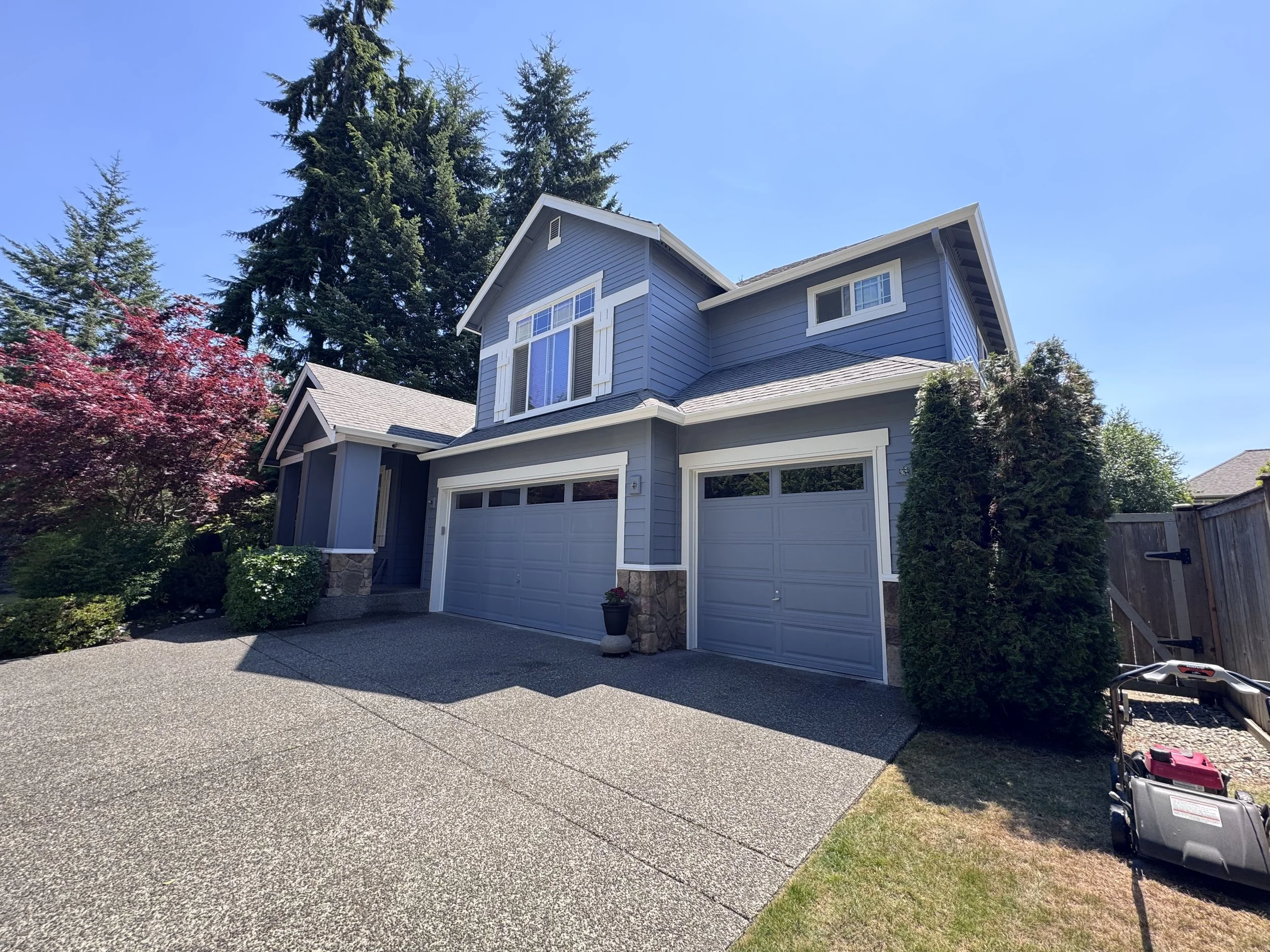 Front view of a light blue, two-story house with a white garage door, surrounded by trees and a small garden. Clear blue sky overhead.