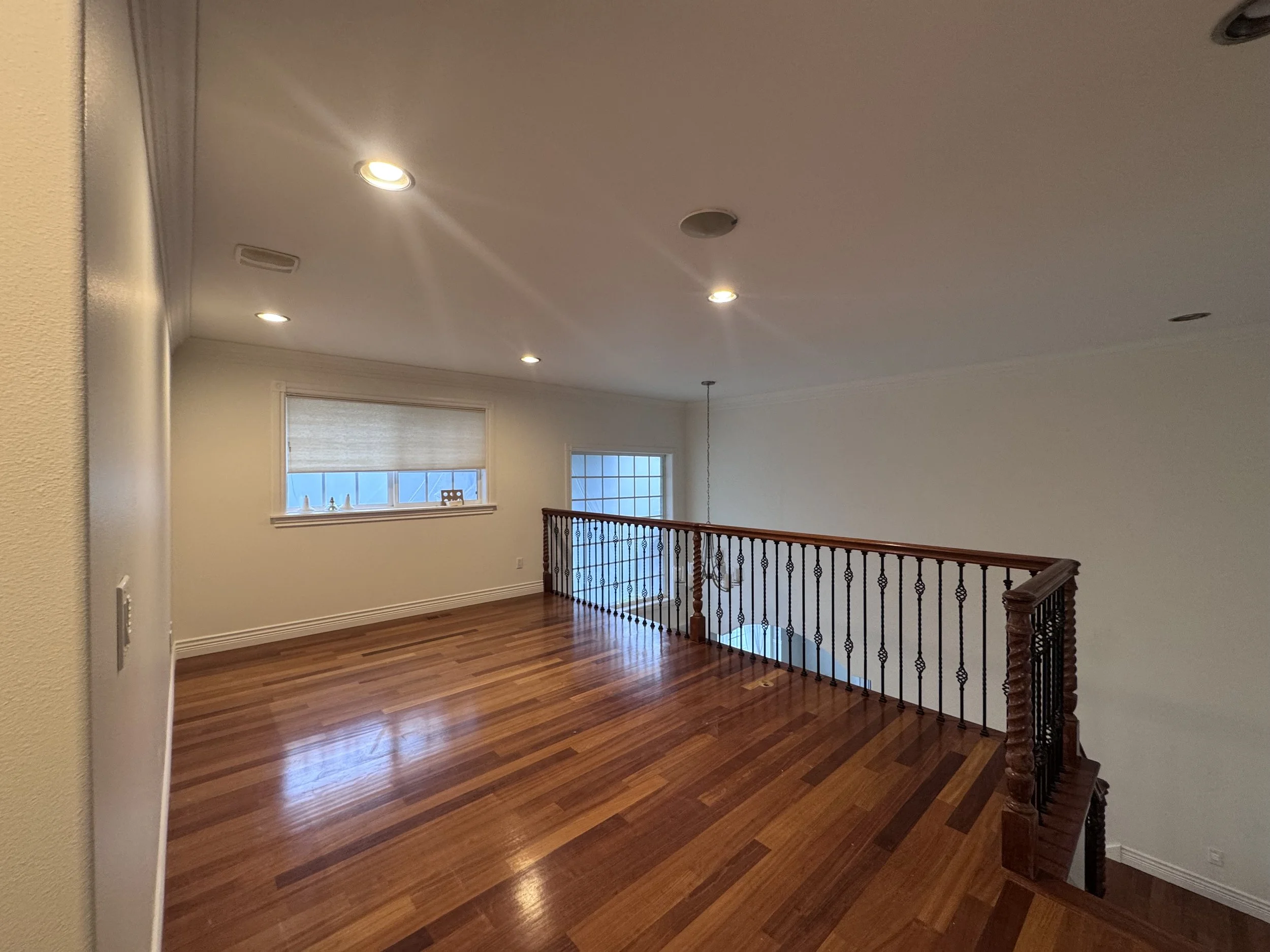 The image shows an open upper-level area of a house with wooden flooring, a staircase with a decorative balustrade, and windows with blinds letting in natural light.
