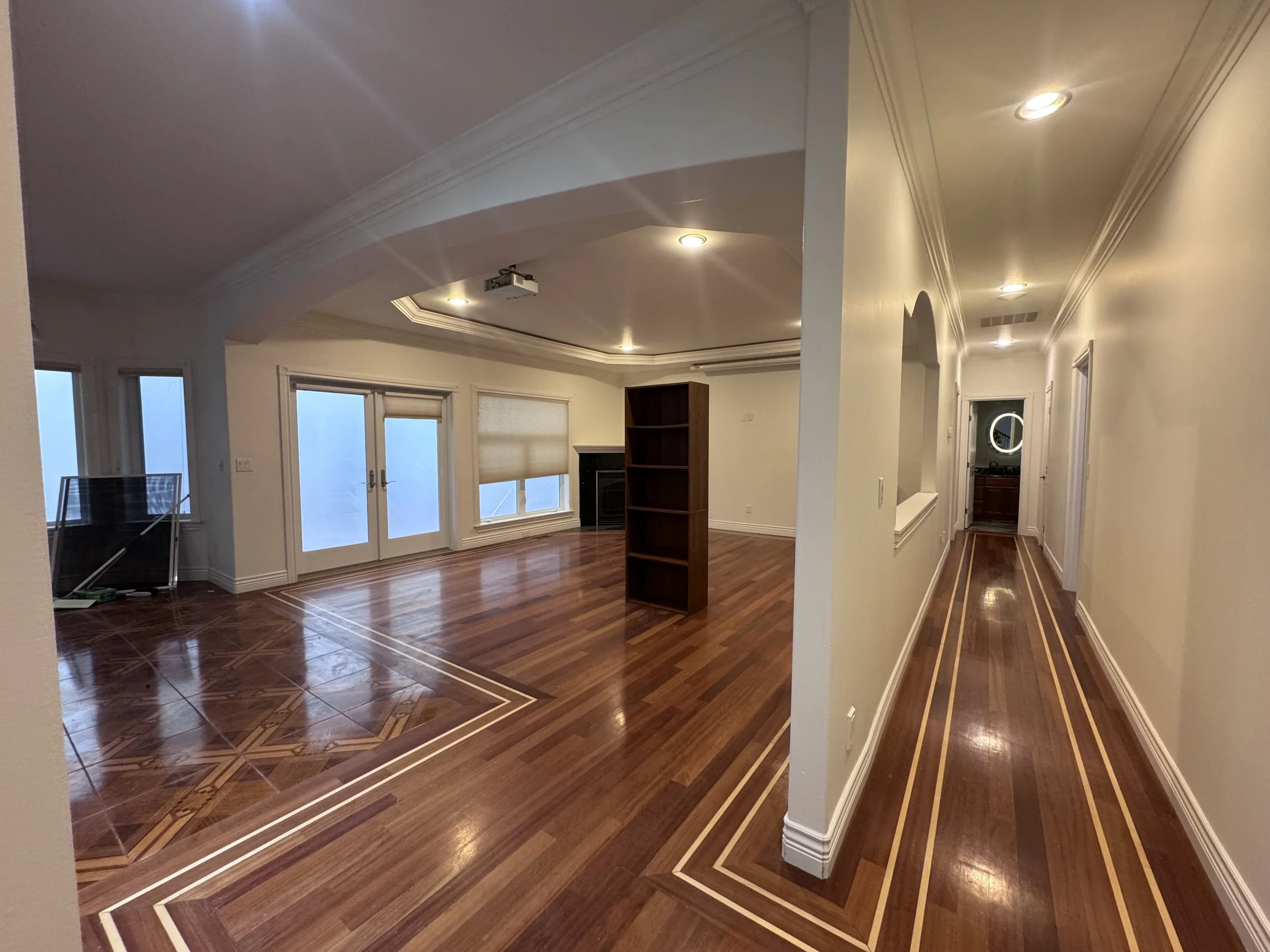 Living room with wood floors, large windows, and a hallway leading to a door with a circular mirror. Recessed lighting and crown molding are visible.