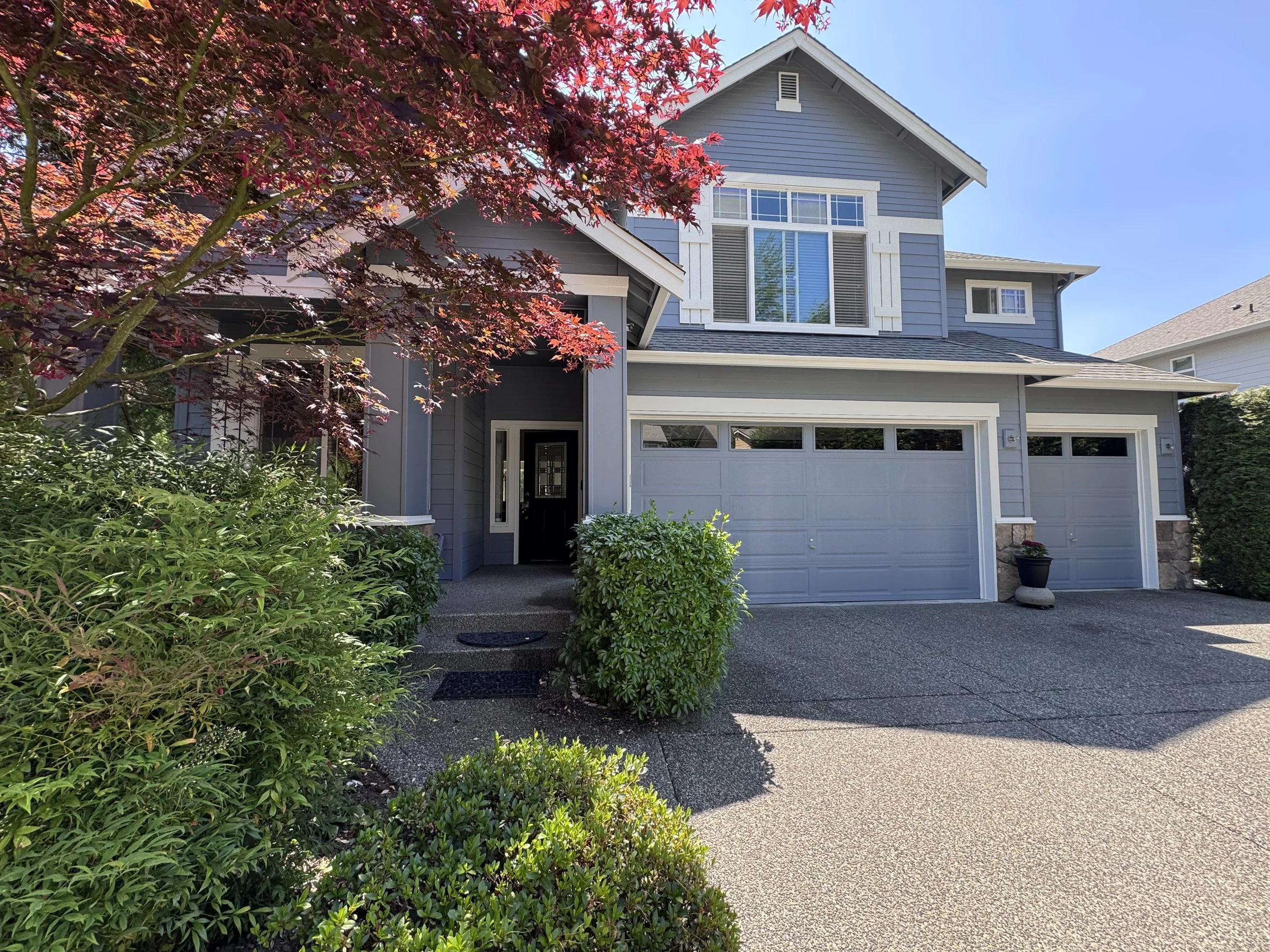 A two-story house with grey siding, white trim, and a garage. A tree with red leaves partially obscures the house. The driveway and walkway are visible, with bushes and plants surrounding the entrance.