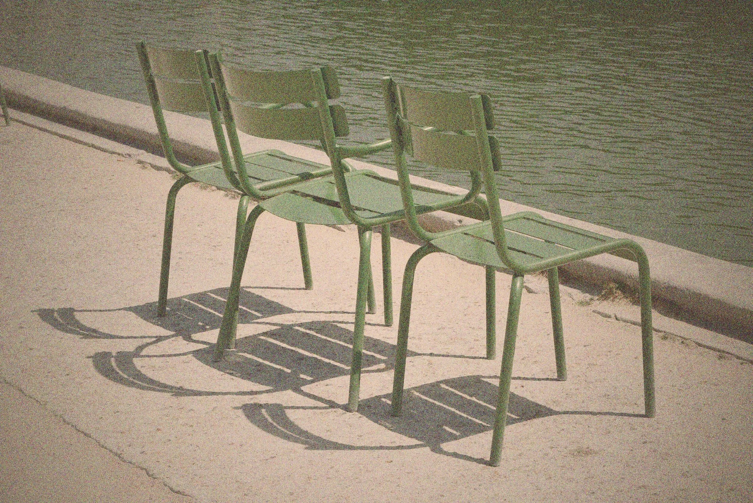 Three green metal chairs on a concrete sidewalk, casting elongated shadows against a dark wall.