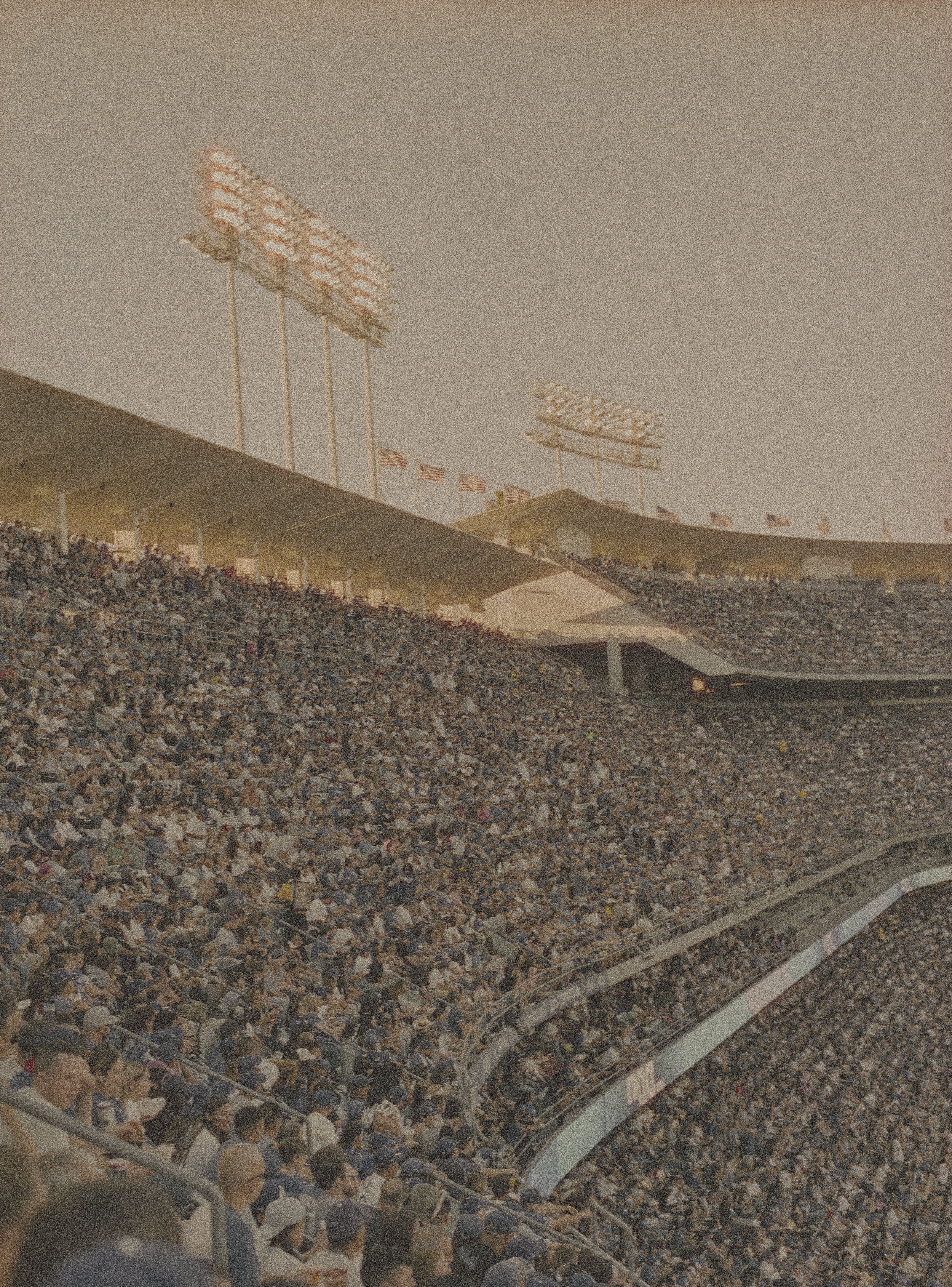 Crowded baseball stadium with fans in bleachers, large floodlights, and American flags.