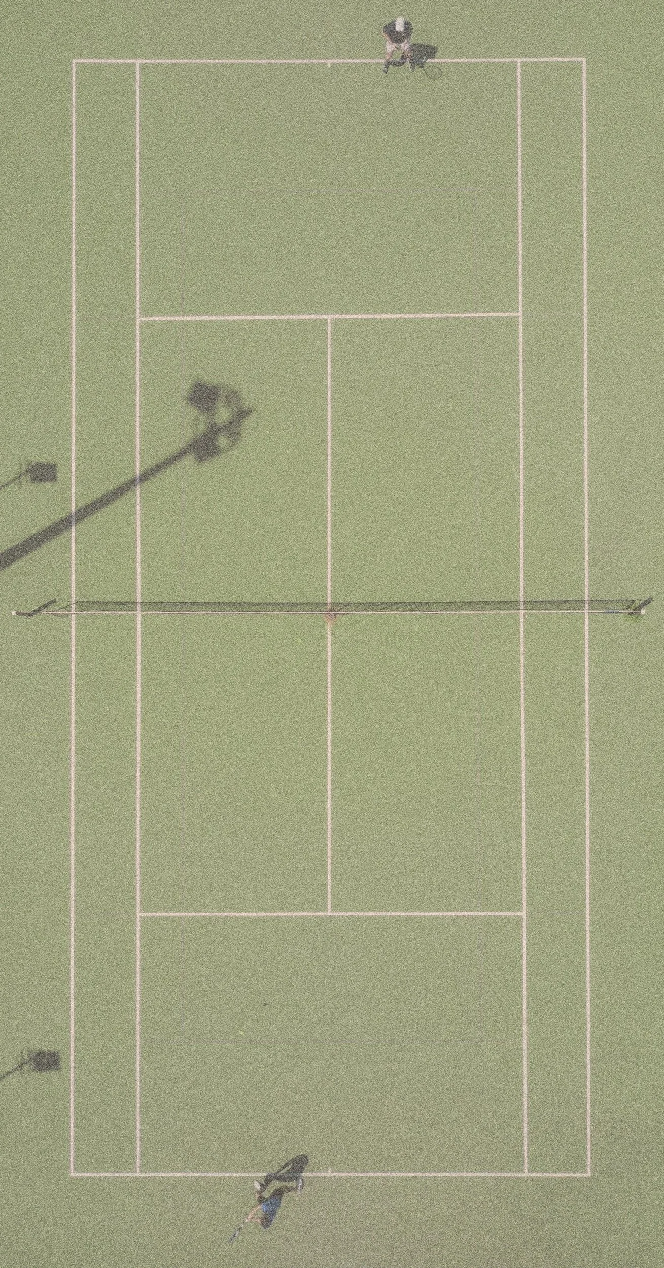 Top-down view of a tennis court with two players, one near each baseline, and a net in the middle, with shadows of the players and court lights cast on the court.