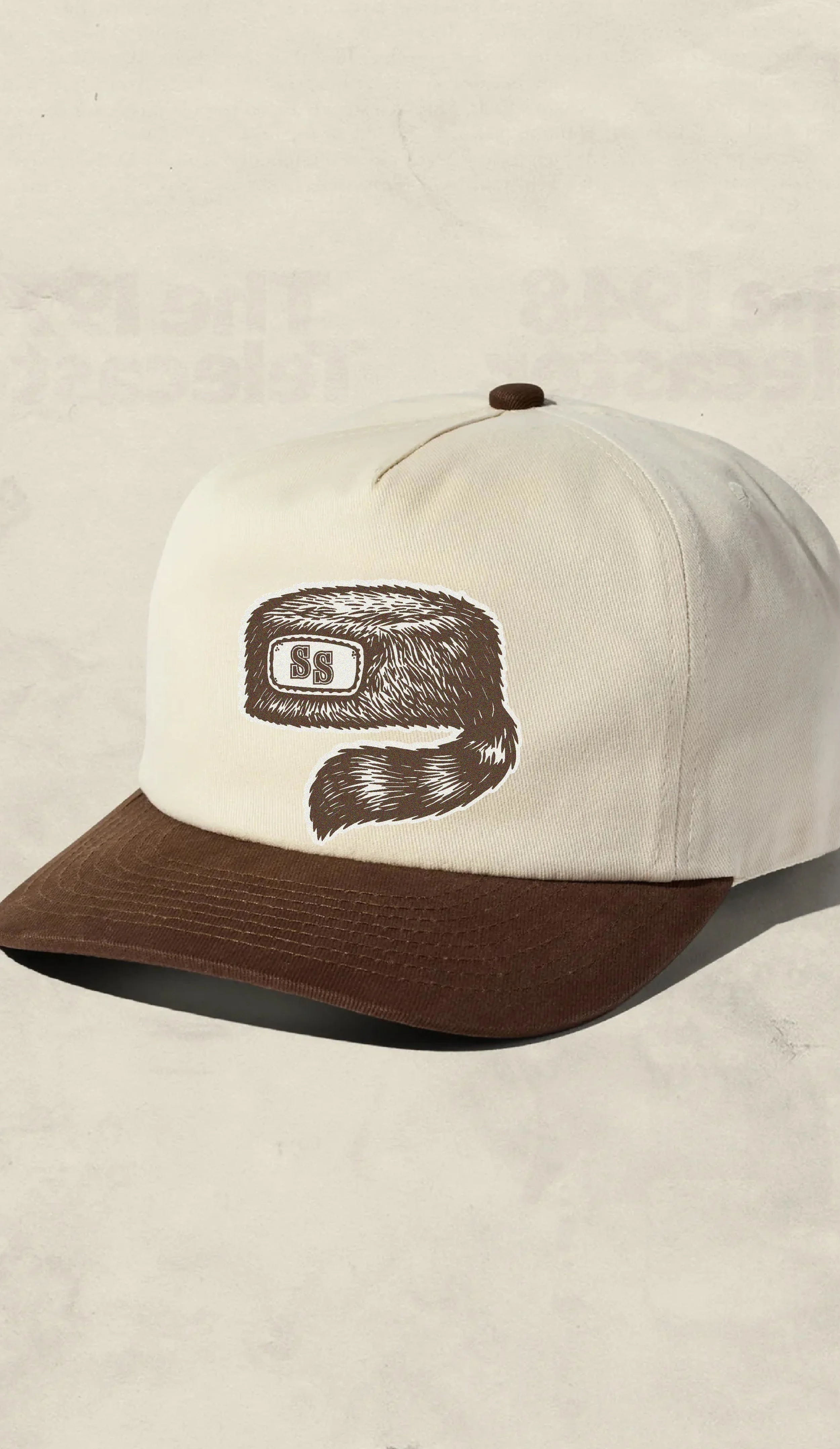Cream-colored baseball cap with a brown brim and a patch of a furry creature's face with glasses and the initials 'SS' on the front.