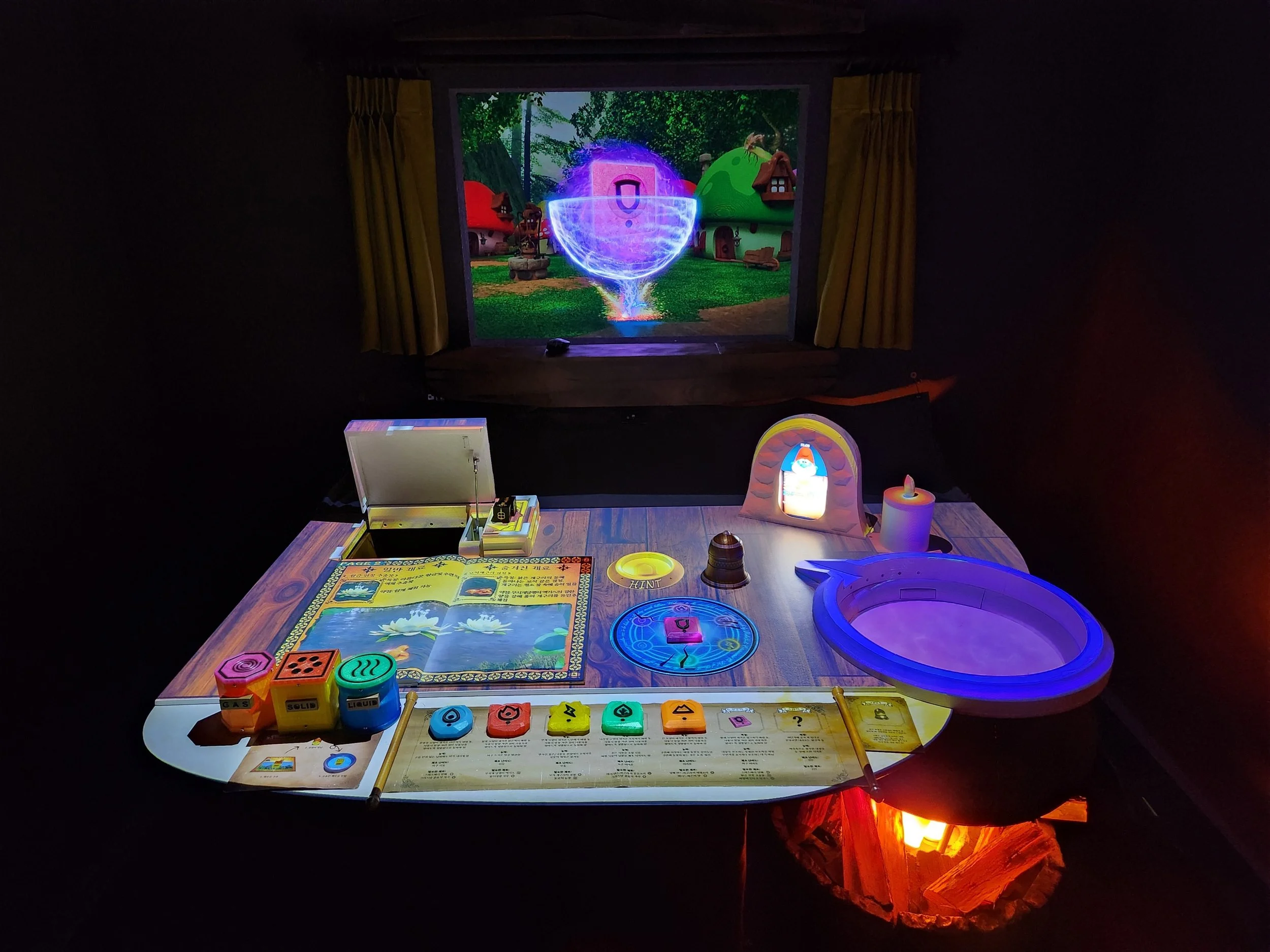 A tabletop setup featuring colorful game pieces, dice, a book, and a board, with a projected animated scene of a fantasy village with houses and a shield emblem in the background.
