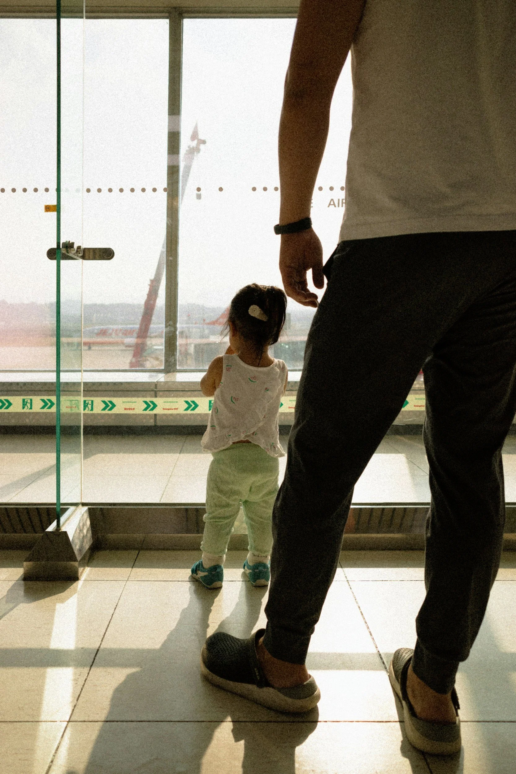 A toddler looking out an airport window with a parent standing beside them, representing the stress and logistics new parents navigate while traveling during the holidays.