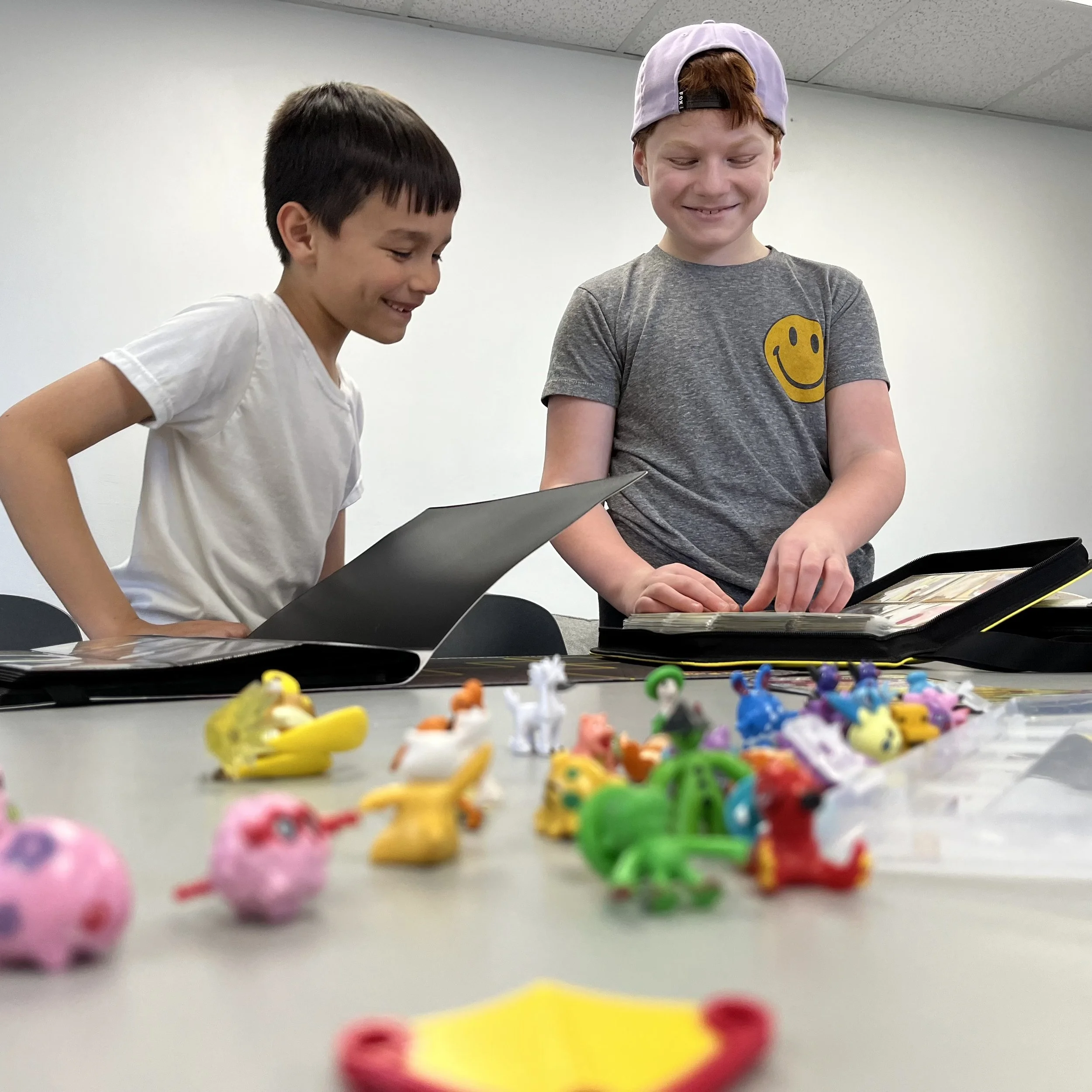 Two young boys are smiling and looking at toy collections in binders on a table.