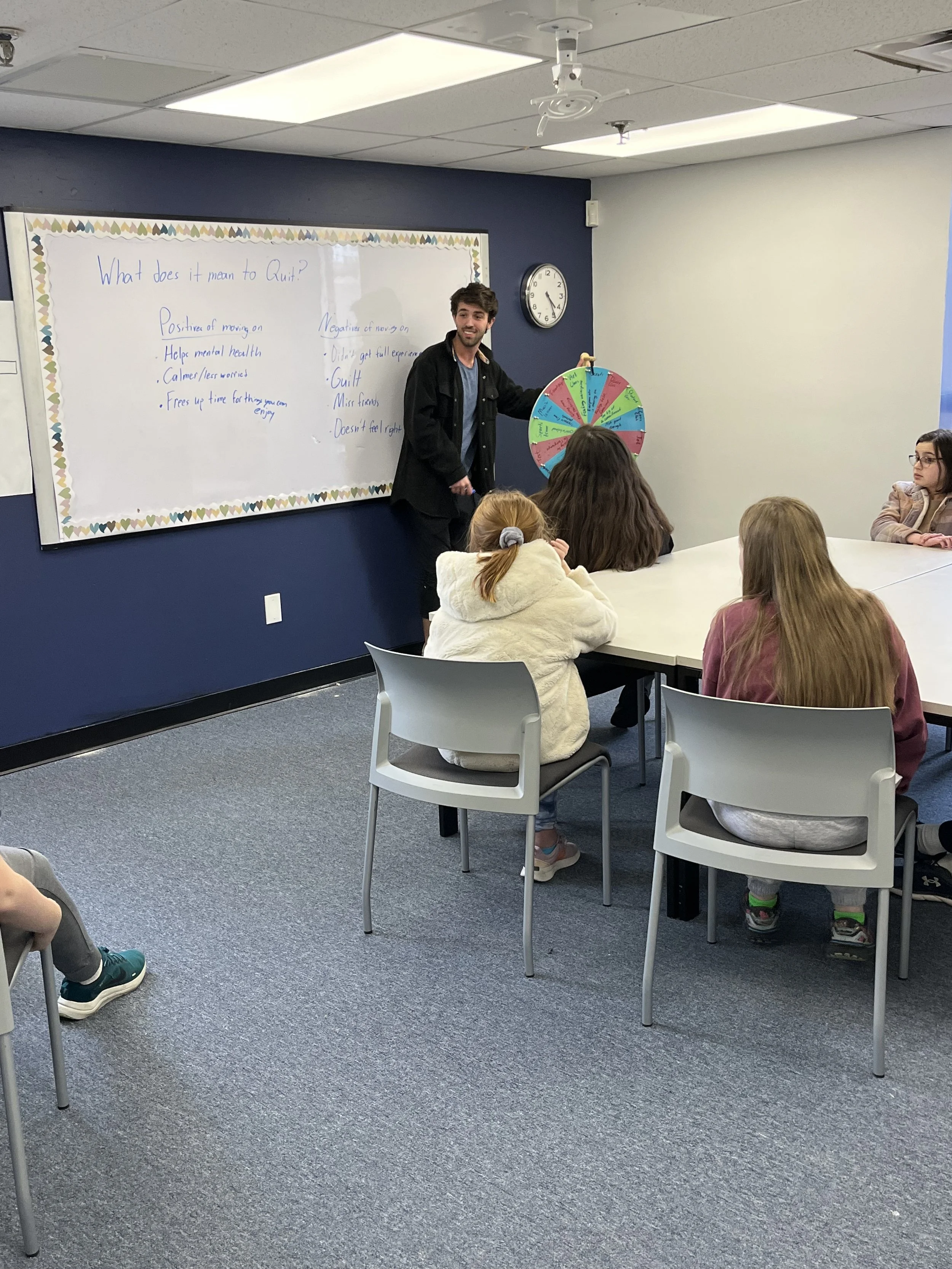 A teacher or speaker standing next to a colorful spinning wheel in front of a classroom of children, with a whiteboard behind him that has notes about quitting. The classroom has gray carpet, white chairs, and a clock on the wall.