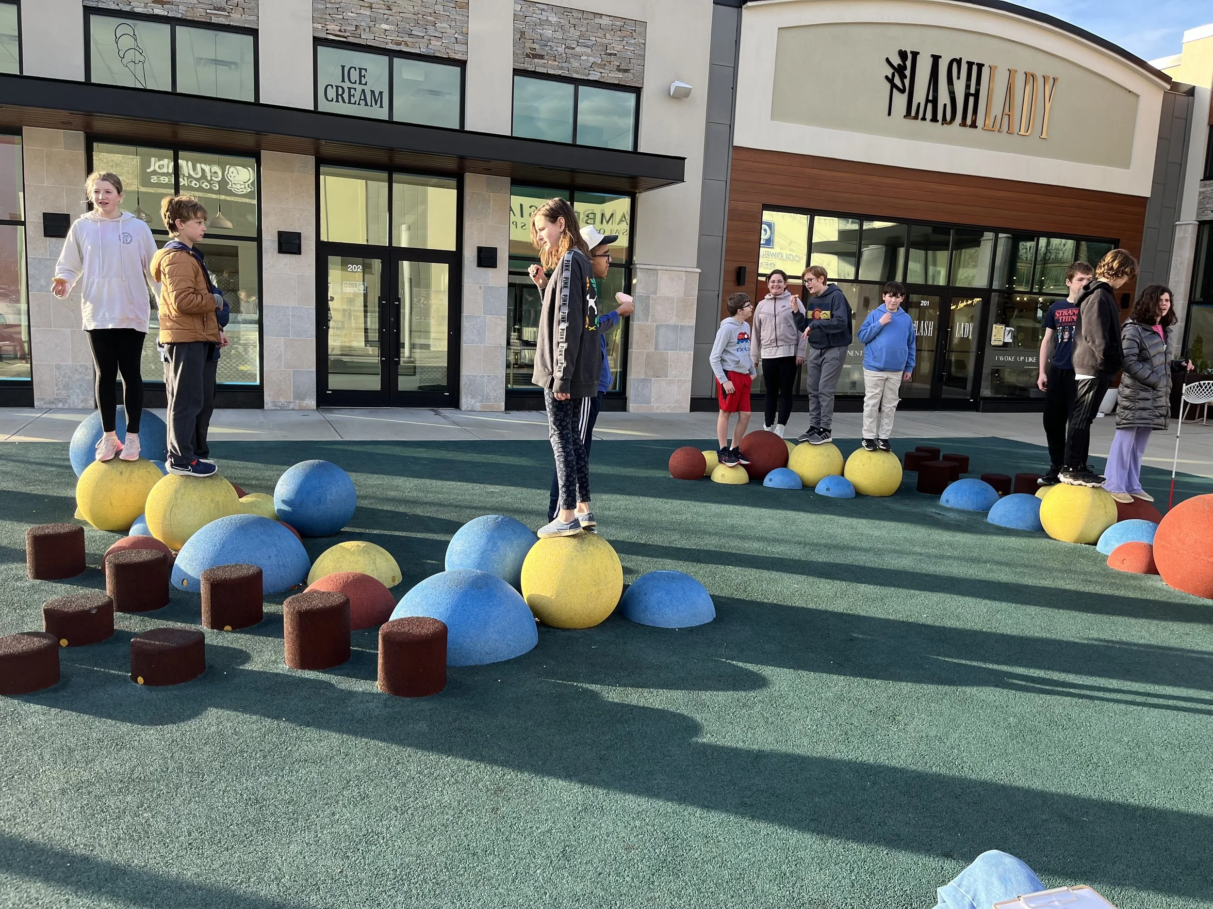 Children playing on colorful, rounded climbing structures outdoors in front of a shopping center.