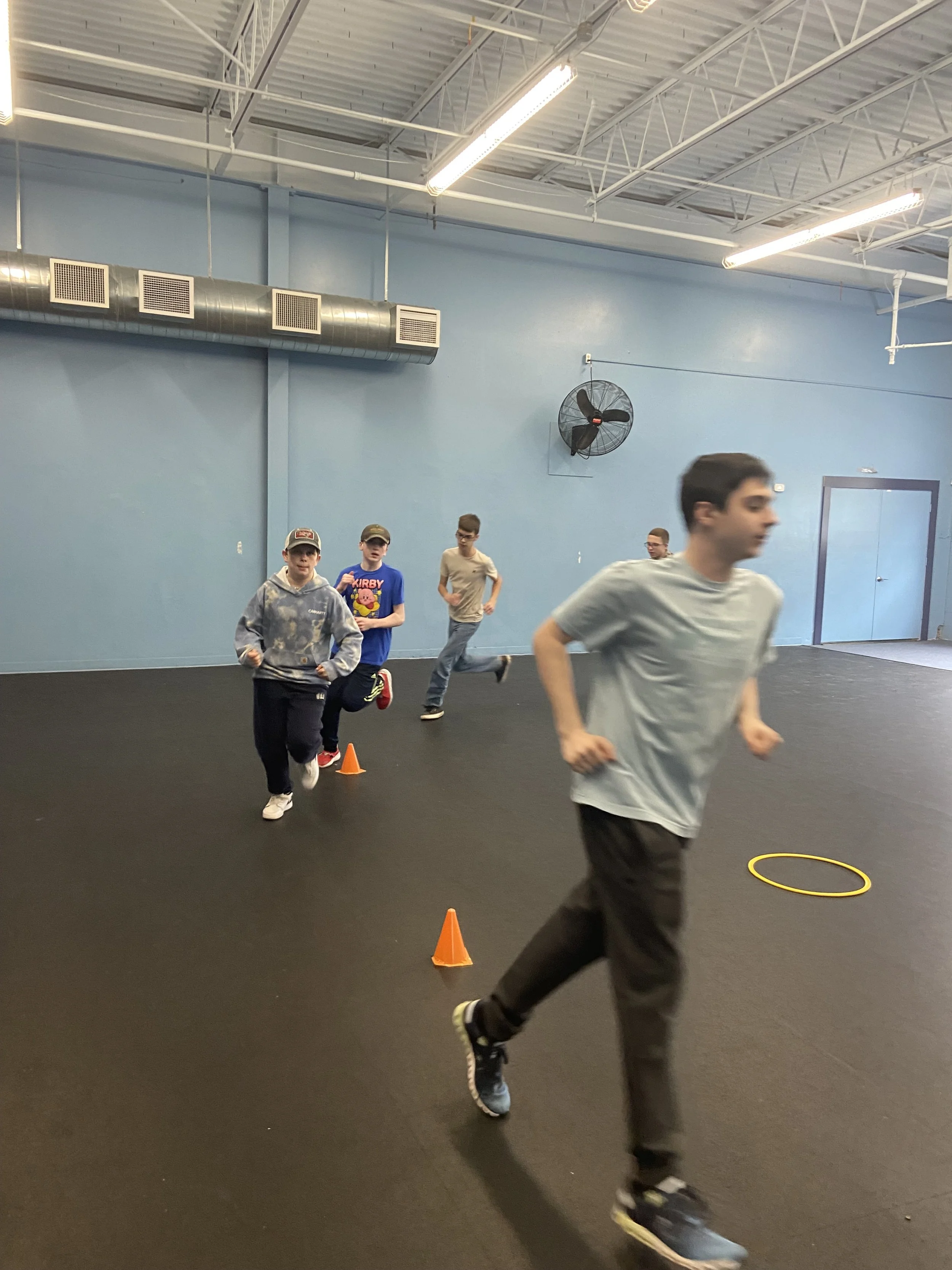 A group of five boys running and playing in an indoor gym, with orange cones and a yellow hoop on the black rubber floor, blue walls, and bright ceiling lights.
