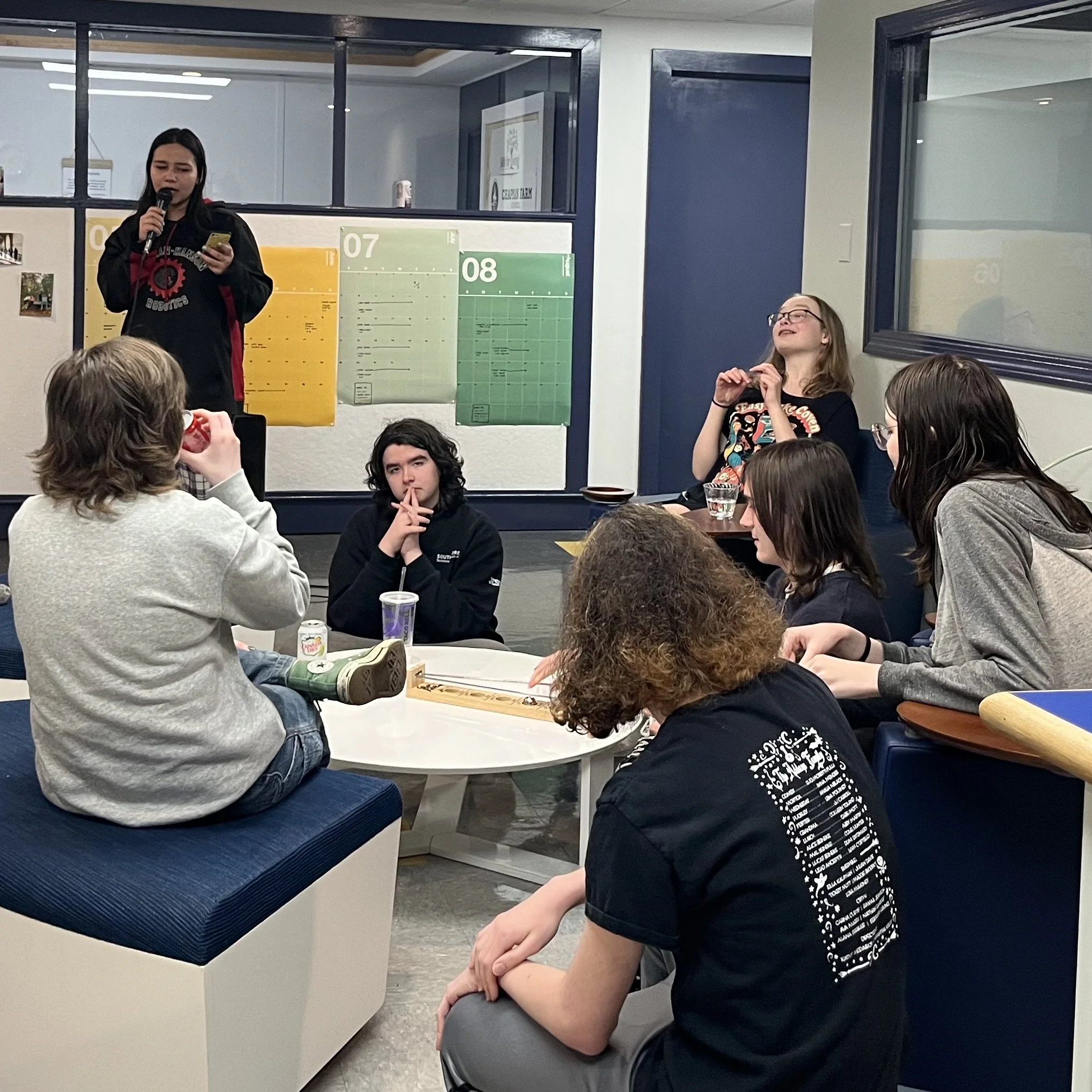 Group of young people gathered around a table with a woman standing and speaking into a microphone in a modern indoor space, with pandas posters on the window behind.