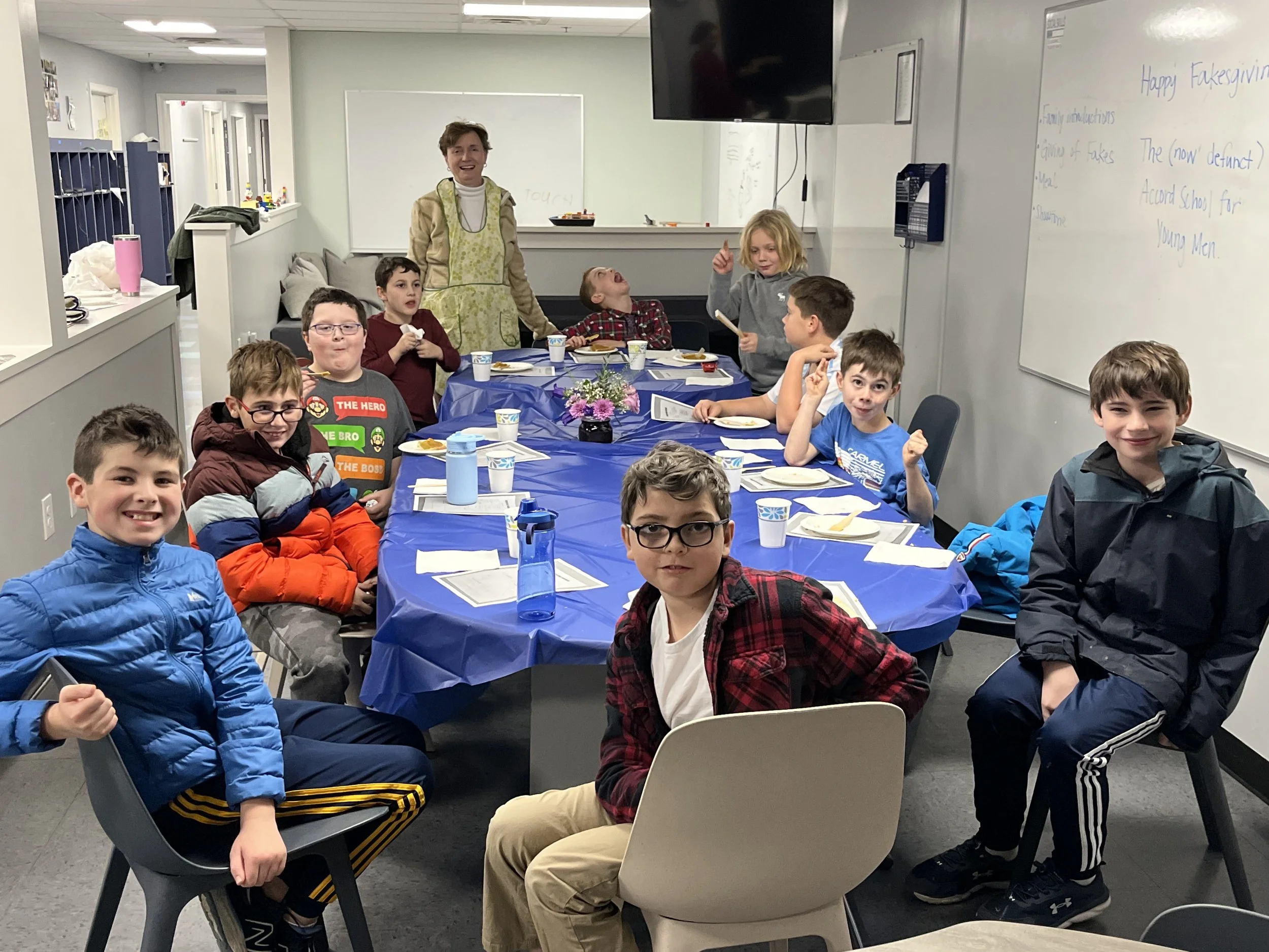 Children and a teacher sitting around a table with blue tablecloth, having a meal in a classroom, celebrating a festive occasion.