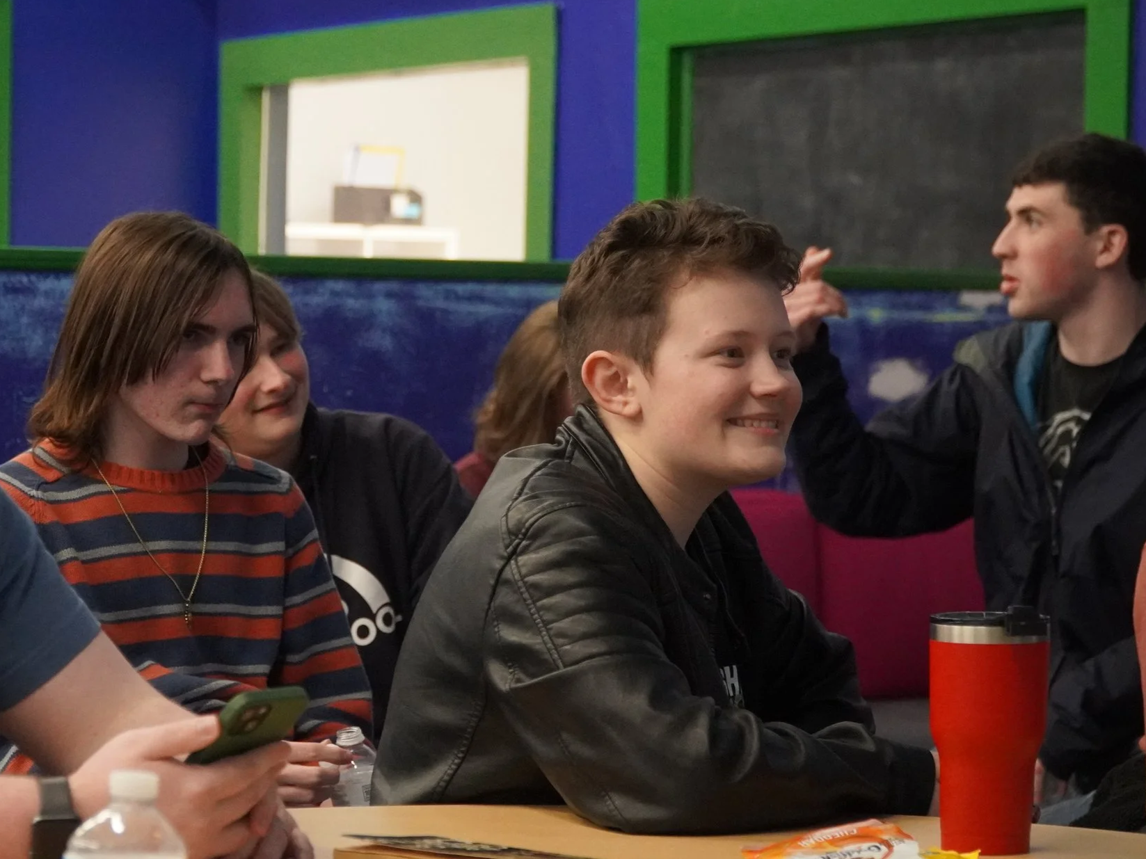 A group of young people sitting at a table in a room with blue walls and green-framed windows, some smiling and chatting, one using a smartphone, with drinks and snacks on the table.