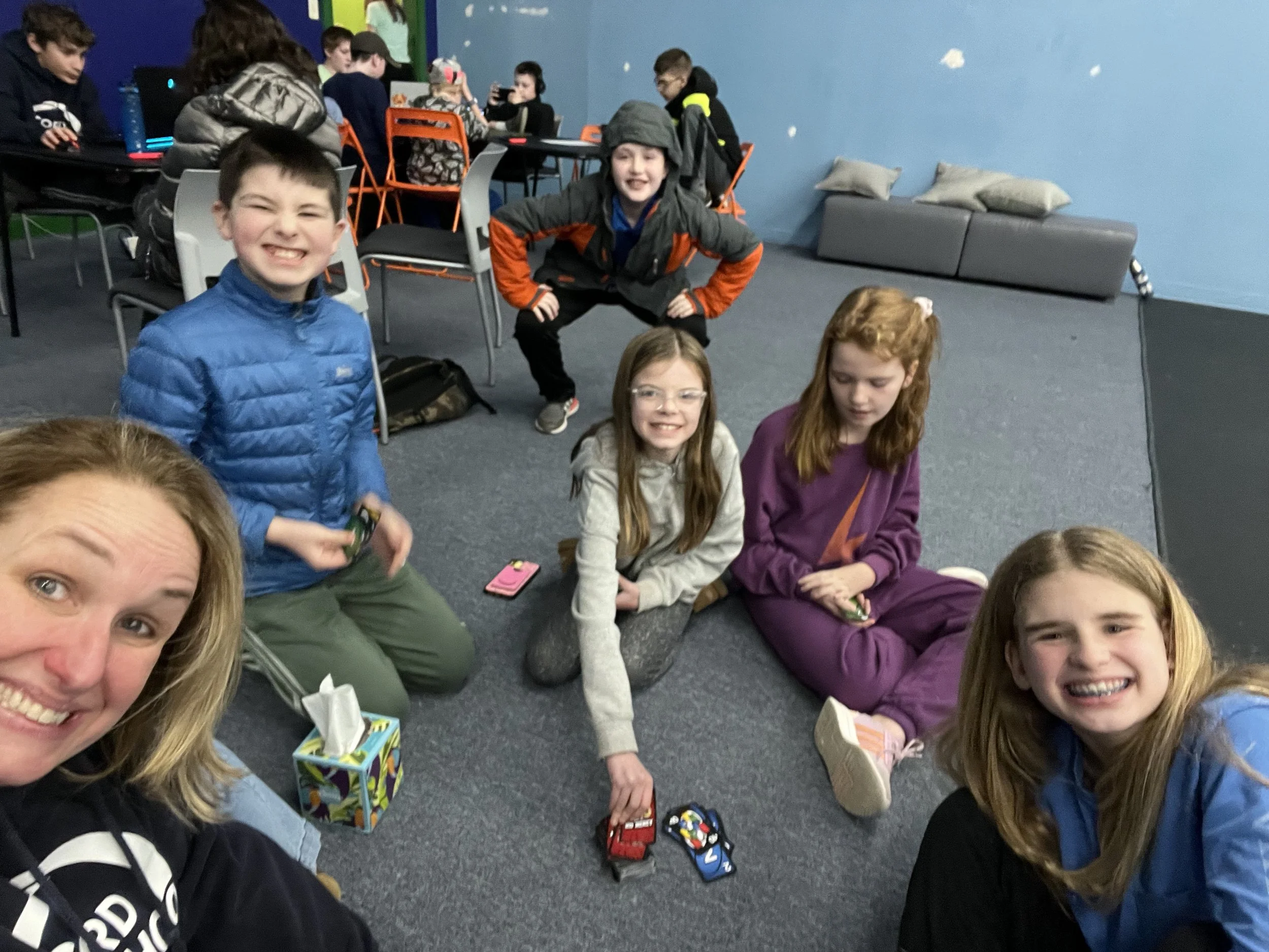 Group of children and a woman sitting and smiling on carpeted floor in a recreational room with blue walls and a gray couch in the background.