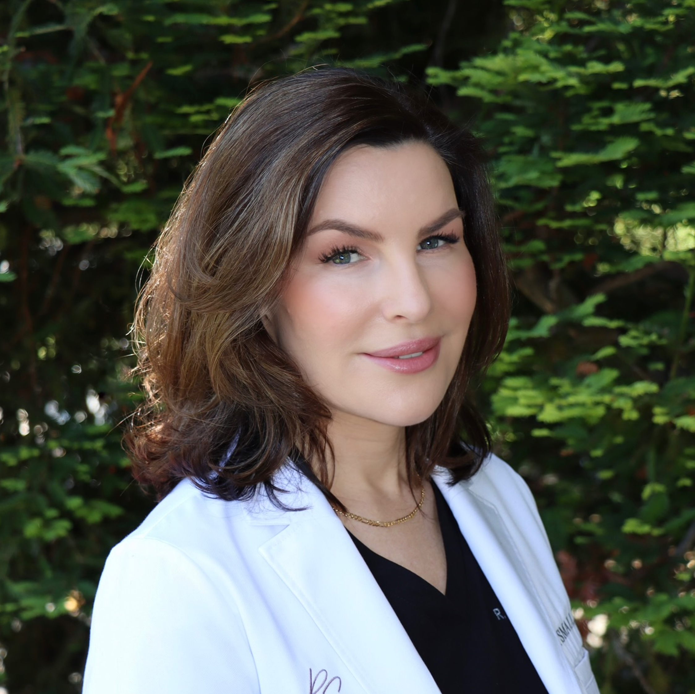Close-up of a woman with brown hair and blue eyes, wearing a white coat and black shirt, standing outdoors against a background of green foliage.