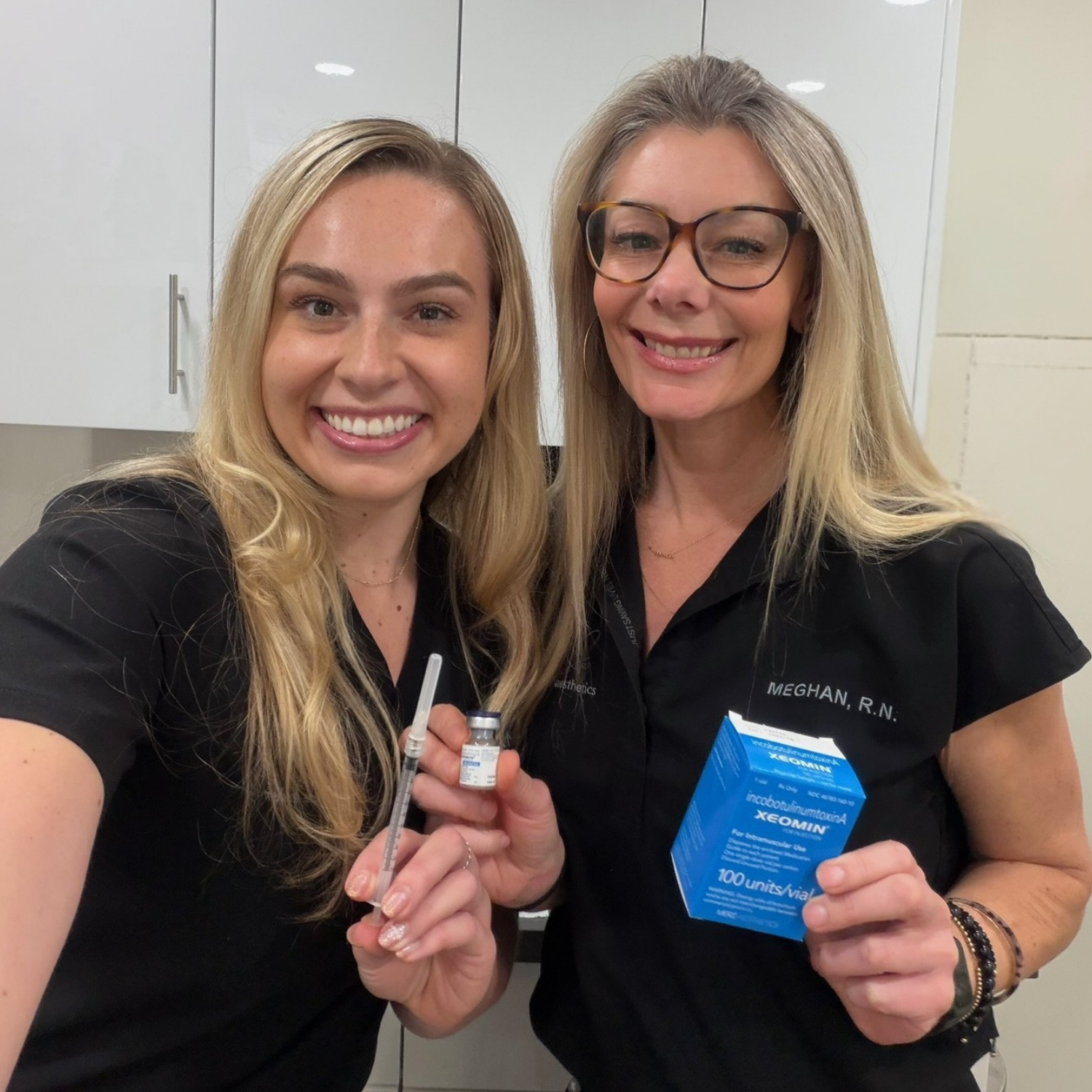 Two women in black scrubs smiling and holding medical supplies, including a syringe, a small vial, and a box of medication, in a clinical setting.