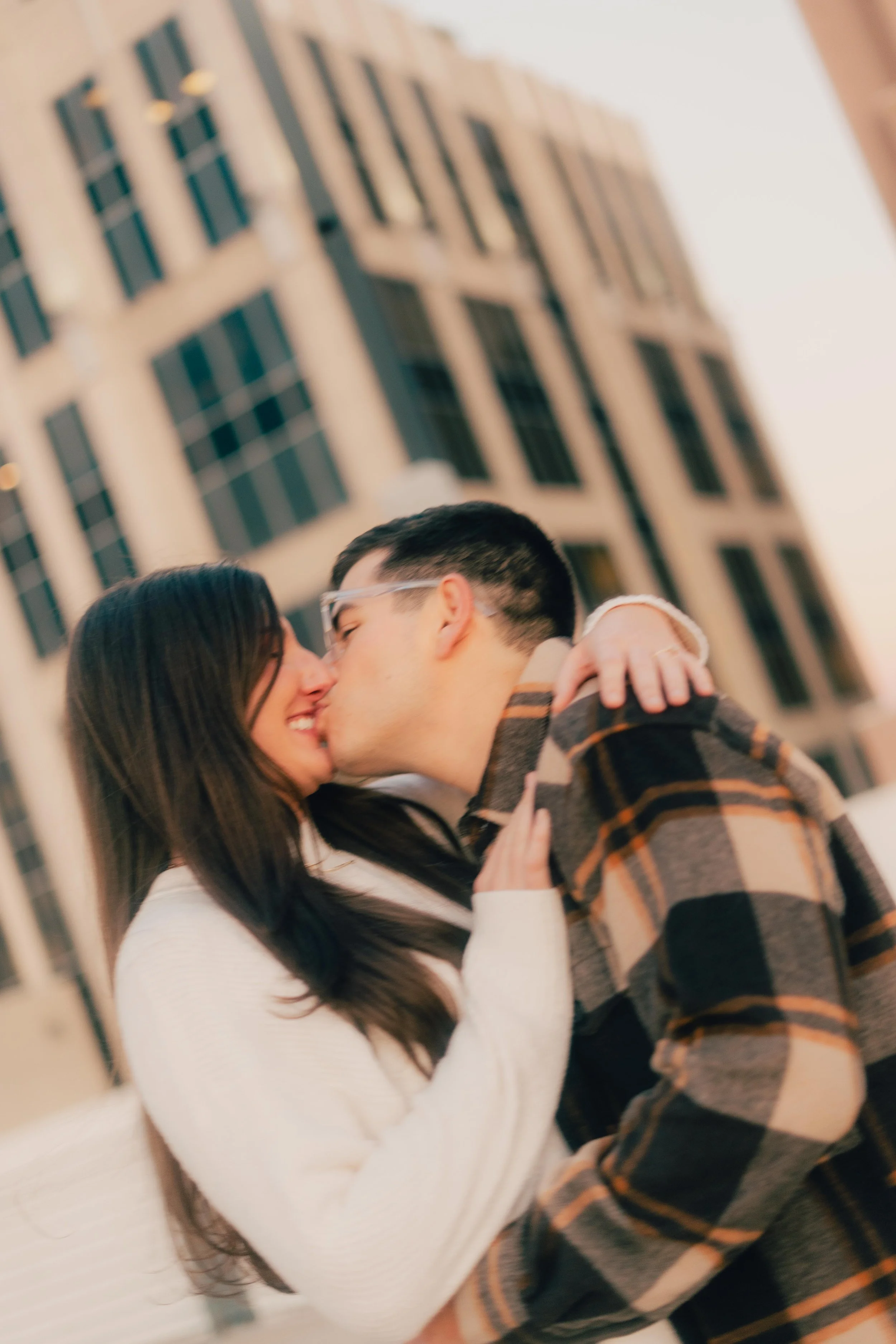 A young couple sharing a kiss outdoors in front of a tall building, with the man wearing glasses and a plaid shirt and the woman with long dark hair wearing a cream sweater.