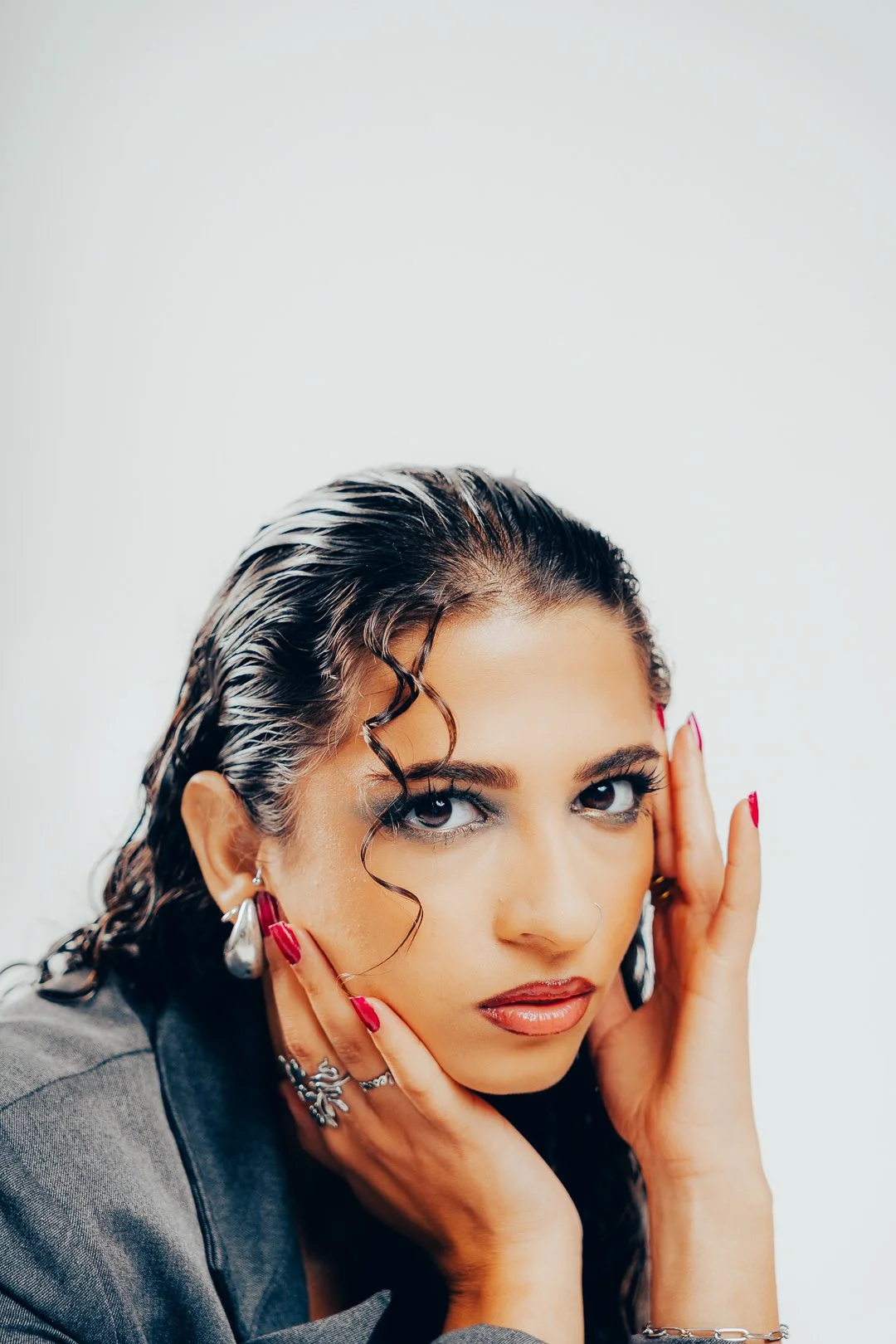 A woman with wet, curly dark hair and light makeup, wearing earrings and jewelry, posing with her hands gently on her face against a plain white background.