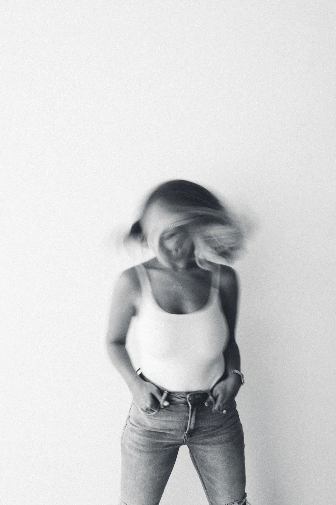 A black-and-white photo of a woman standing against a plain wall, with her head tilted downward and her hair in motion, wearing a tank top and jeans.