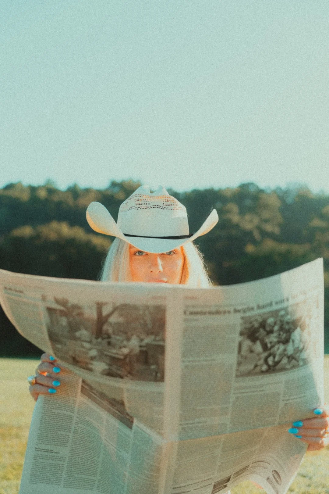 A woman with blonde hair wearing a cowboy hat is reading a newspaper outdoors with trees in the background.