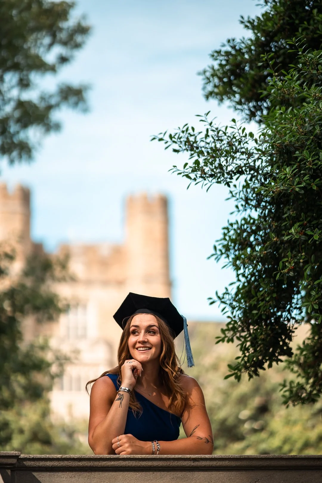 A young woman wearing a graduation cap sits outdoors, smiling and resting her chin on her hand, with a historic building in the background.