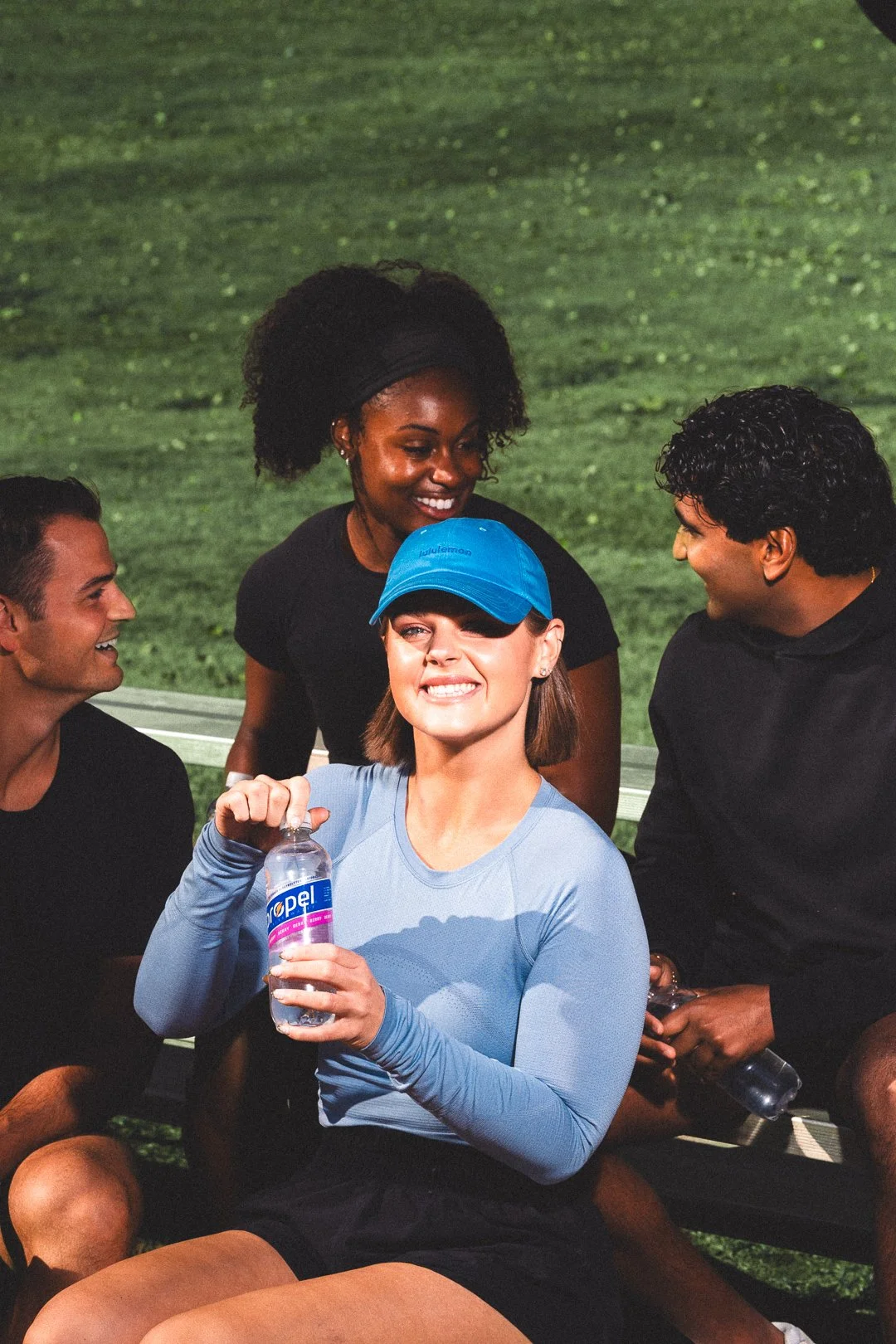 Four young adults sitting on a park bench smiling and socializing, one woman in a blue cap holding a water bottle, others engaging in conversation, outdoors on a grassy field.