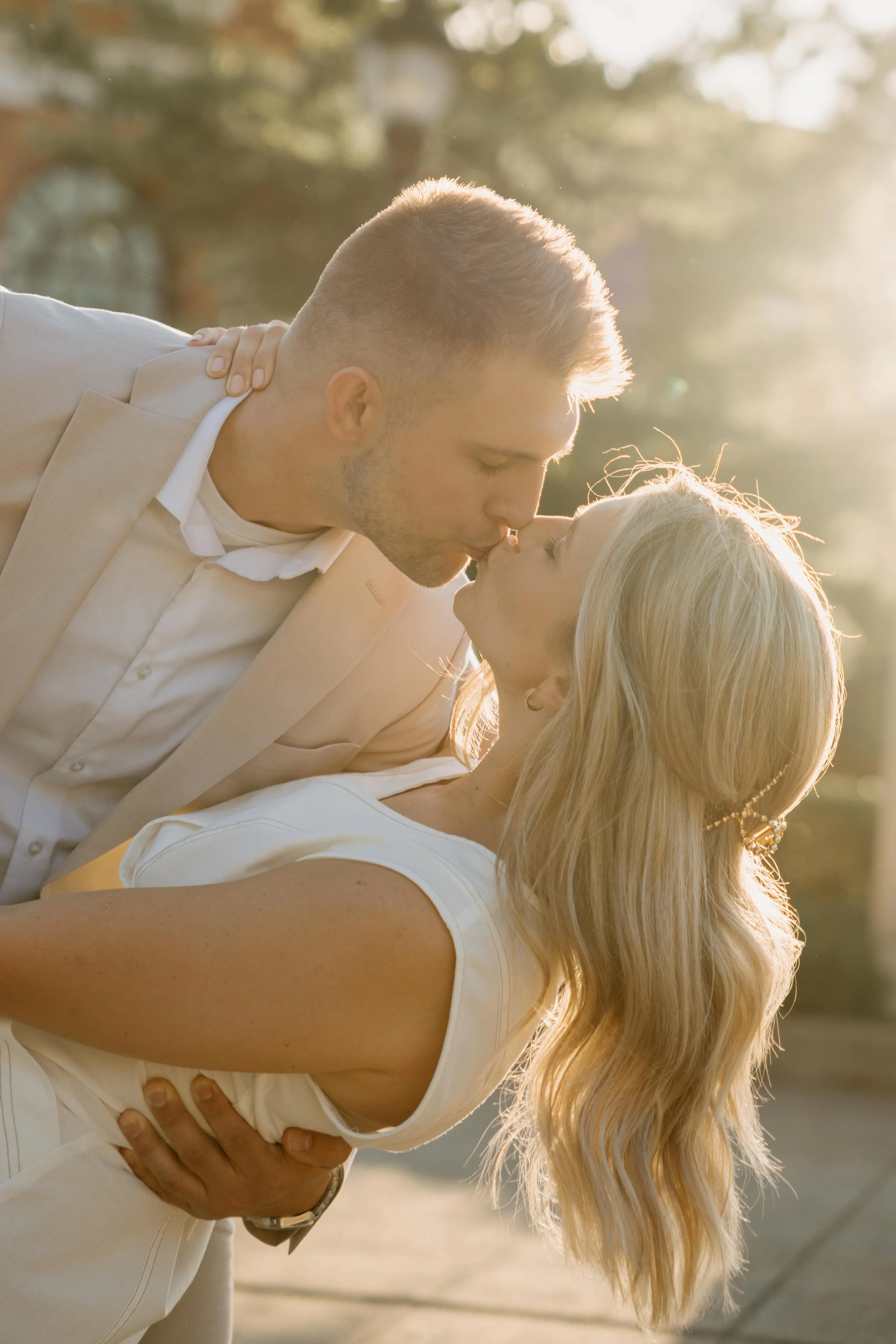 A couple shares a kiss outdoors in bright sunlight, with trees in the background.