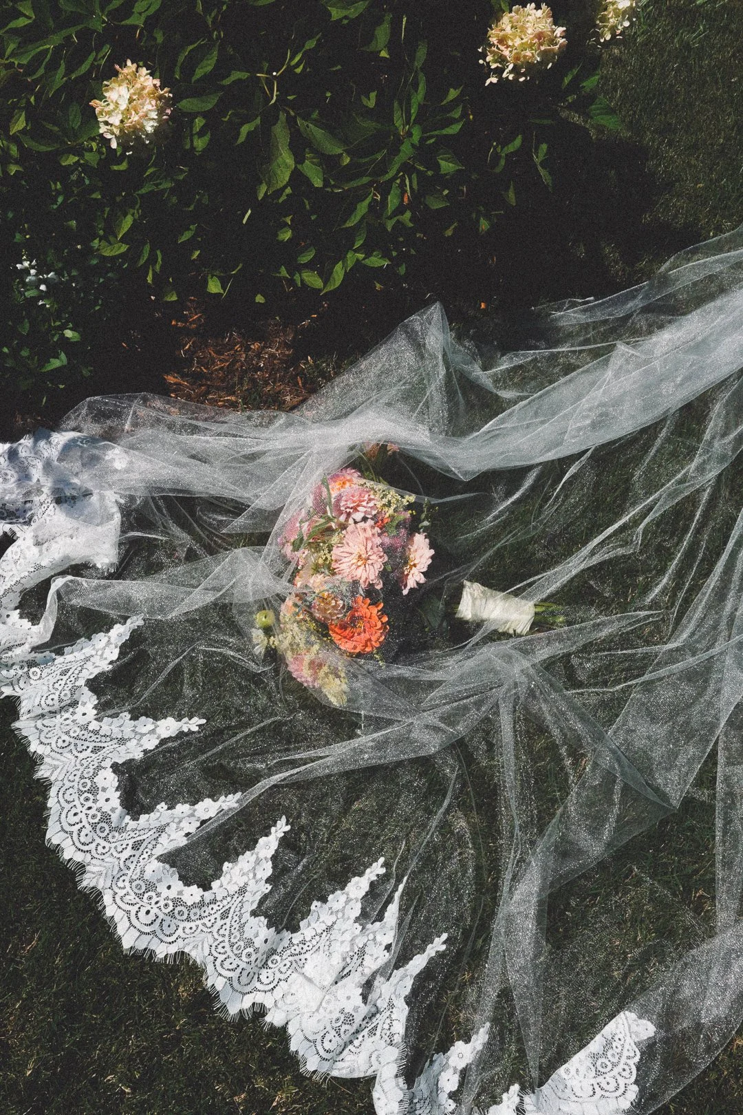 A bouquet of pink, orange, and purple flowers wrapped in paper, lying on a lace-trimmed lace veil with white lace patterns, on the ground next to a bush with pink flowers, outdoors.