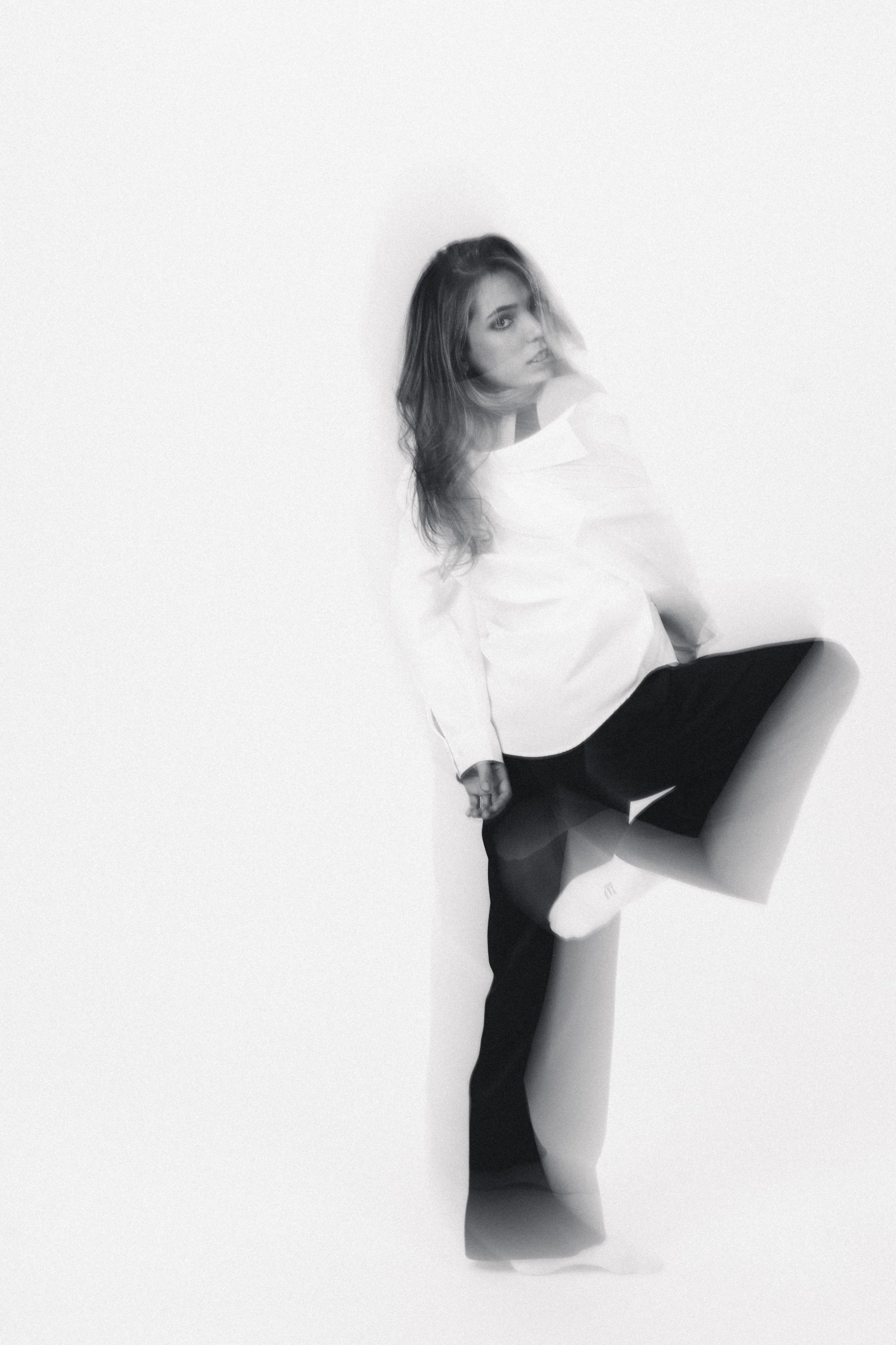 Black and white photo of a woman with long hair, sitting against a plain white background. The photo has a double exposure effect, creating a layered look with a dreamy, artistic feel.
