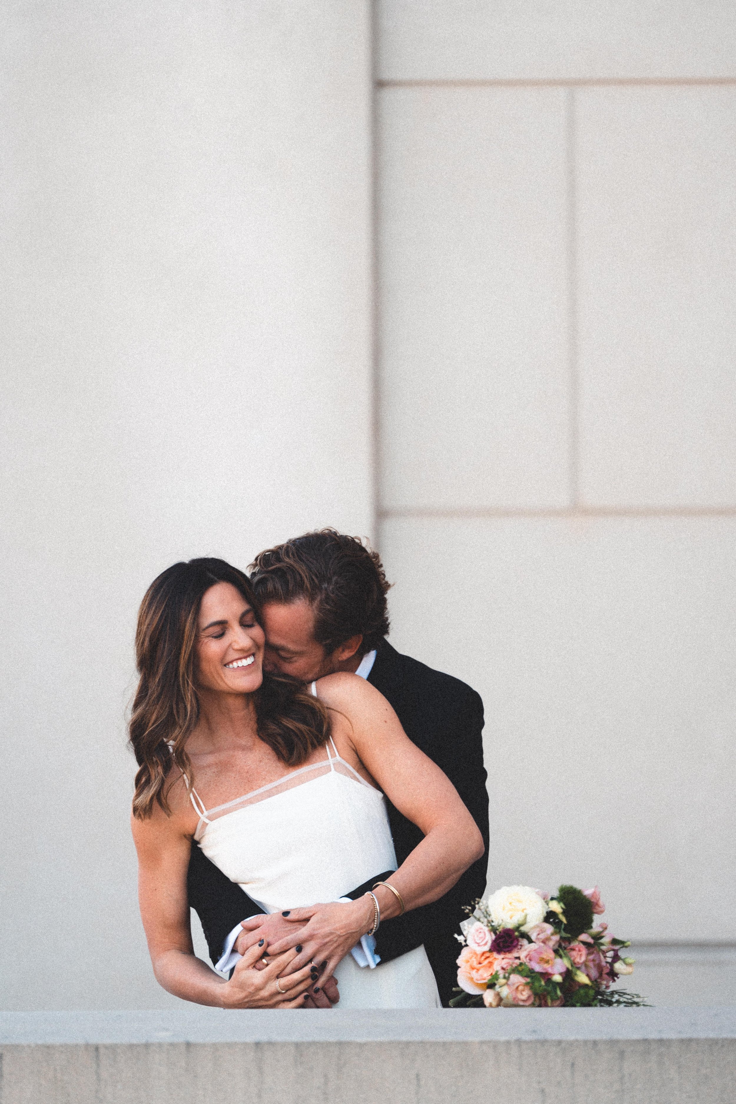 A man and woman smiling and hugging on a rooftop, with a bouquet of roses and flowers on the counter in front of them.