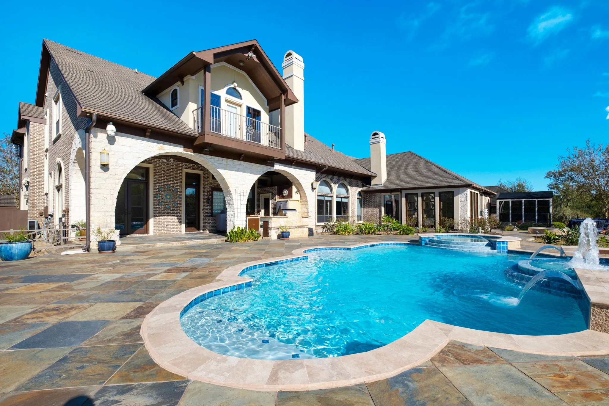 Backyard with a swimming pool in front of a large house with arched windows and a balcony, under a bright blue sky.